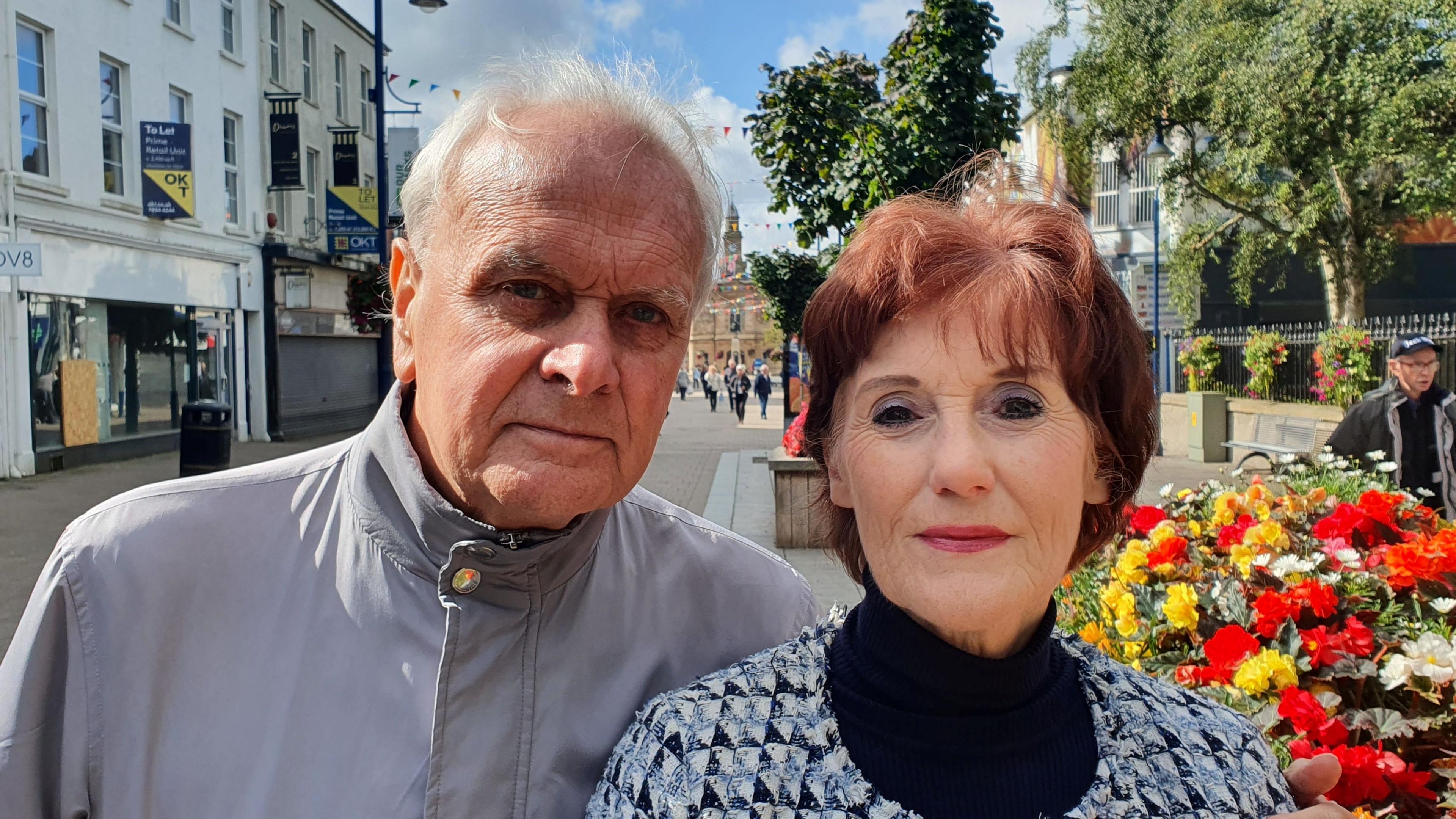 Don McCart and Angela McLaughlin stand together in Coleraine town centre. He is wearing a grey jacket and has his arm around Angela, who is wearing a black top , with a checked jacket. Pedestrians can be seen in the background.