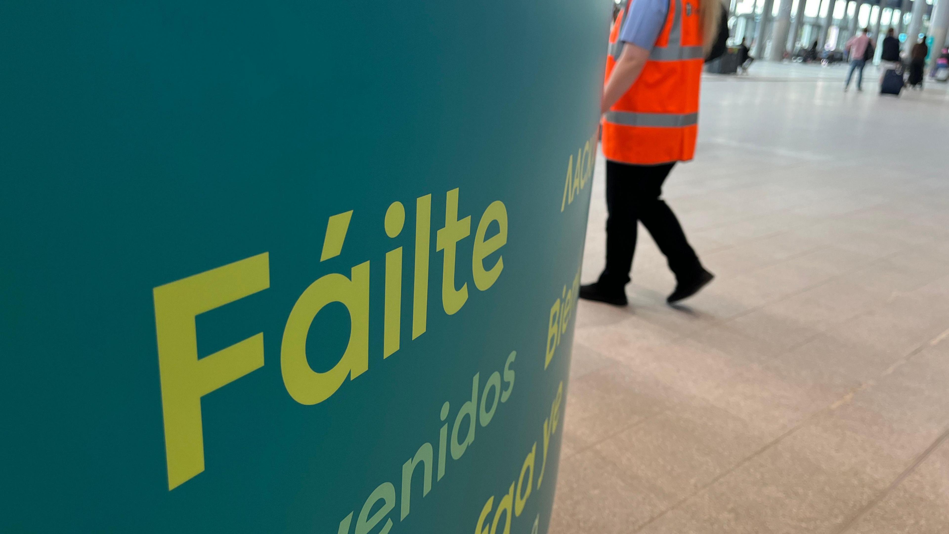 A green sign saying "Fáilte", Irish for "welcome", in the Grand Central Station, with a Translink employee in an orange fluorescent jacket behind. 