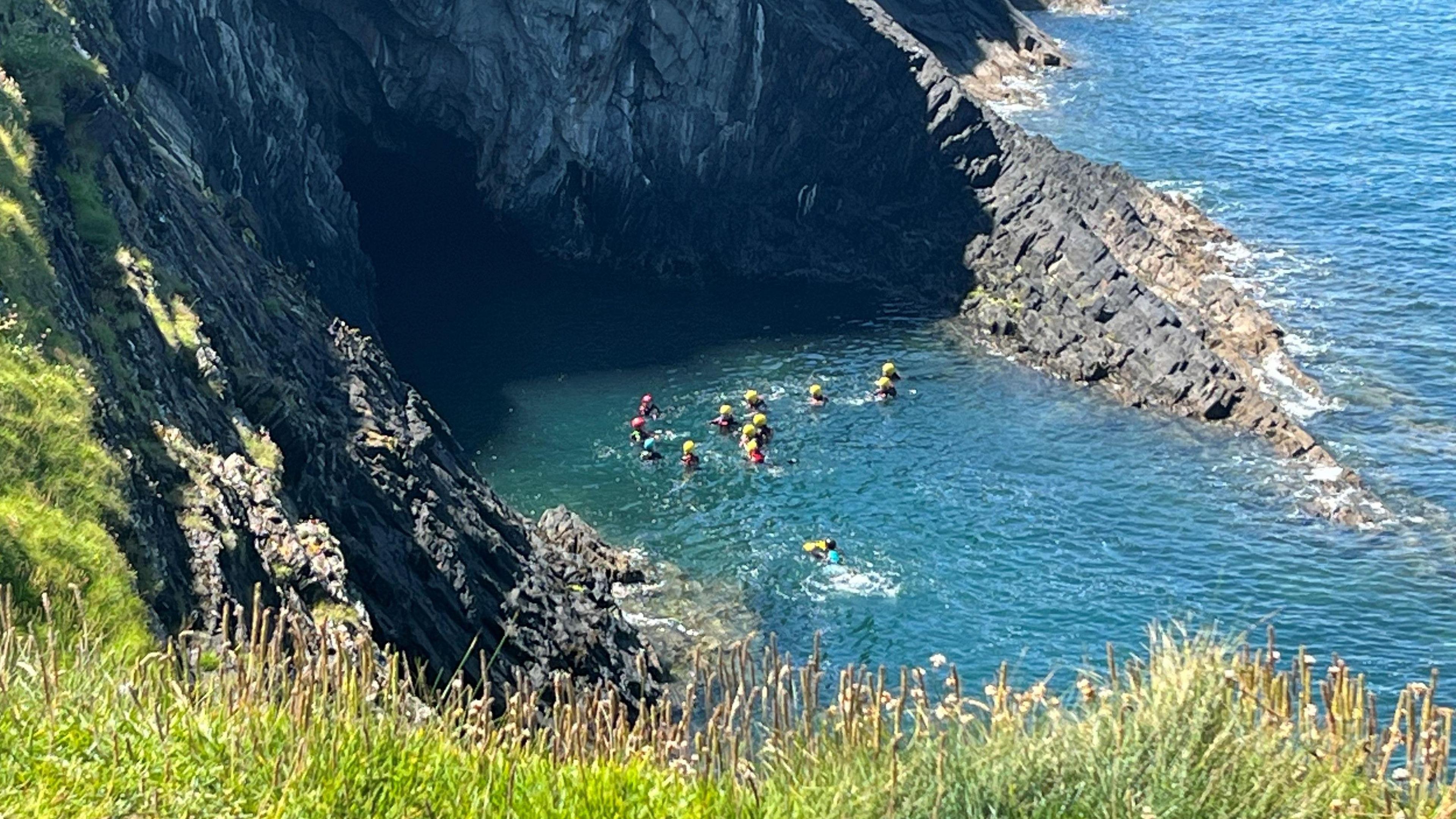 A coasteering group at Ceibwr Bay, the sea is turquoise and the black and grey rocks are jutting out of the sea