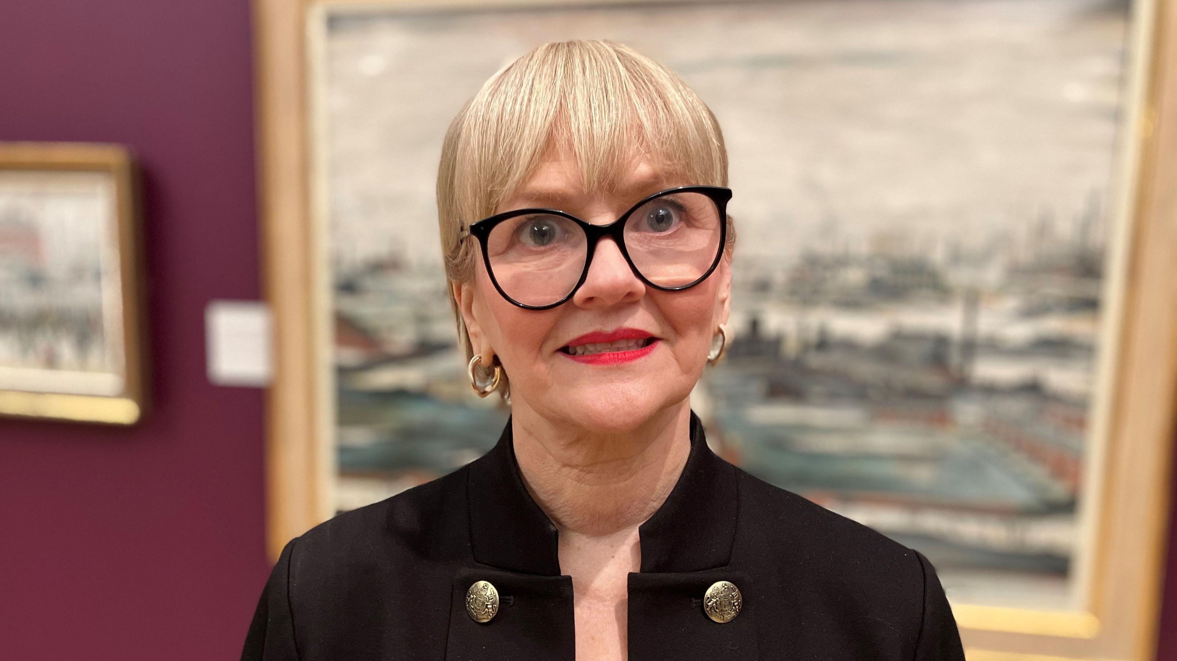 The Chief Executive of The Lowry Julia Fawcett, who has short crop blonde hair and black glasses, smiles for a photograph in front of an LS Lowry painting in an art gallery. 