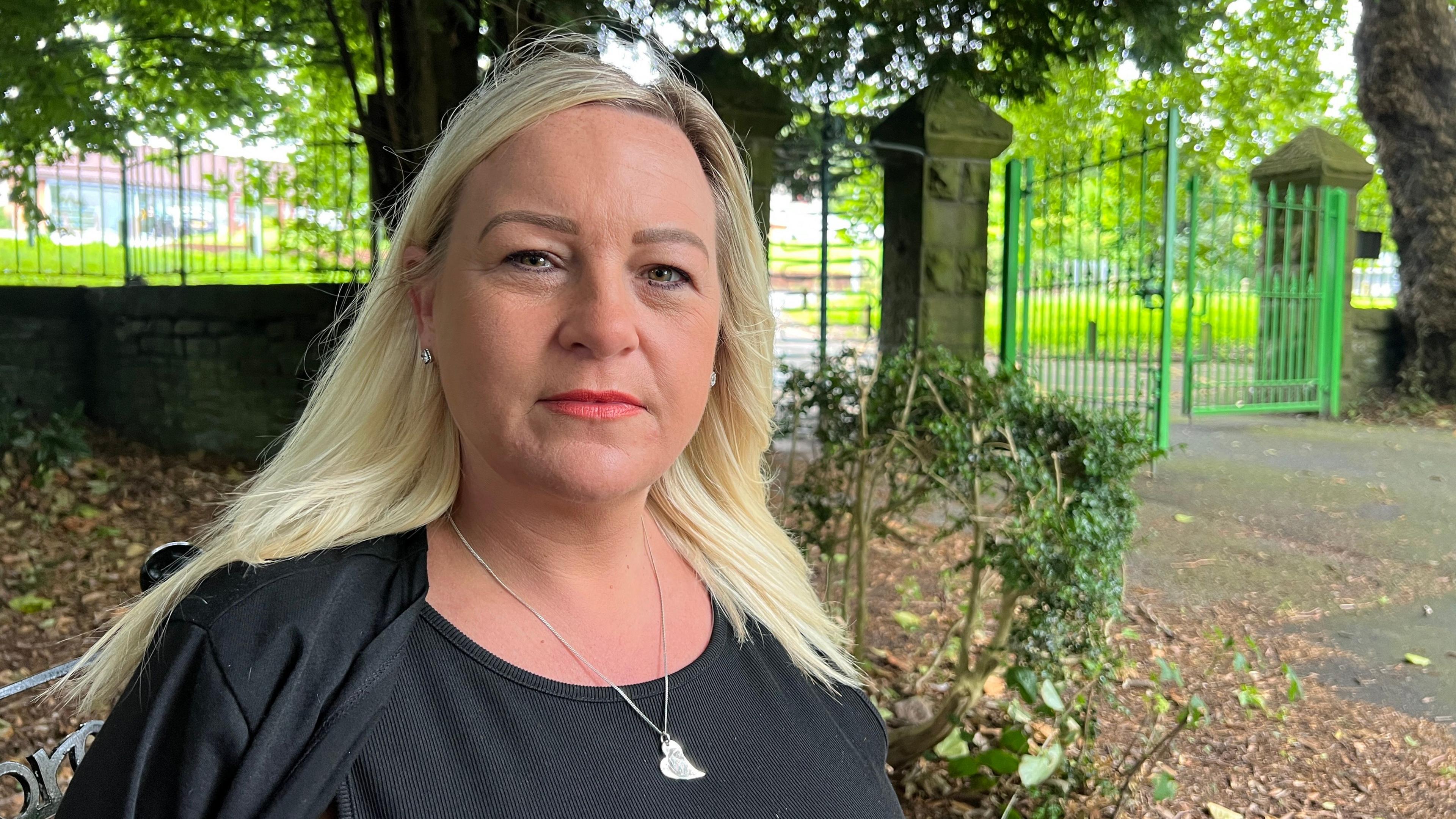 Zoe Day looks straight at the camera, she is sitting in a black iron park bench in her home town of Llansamlet. She has long blonde hair and is wearing a black top.