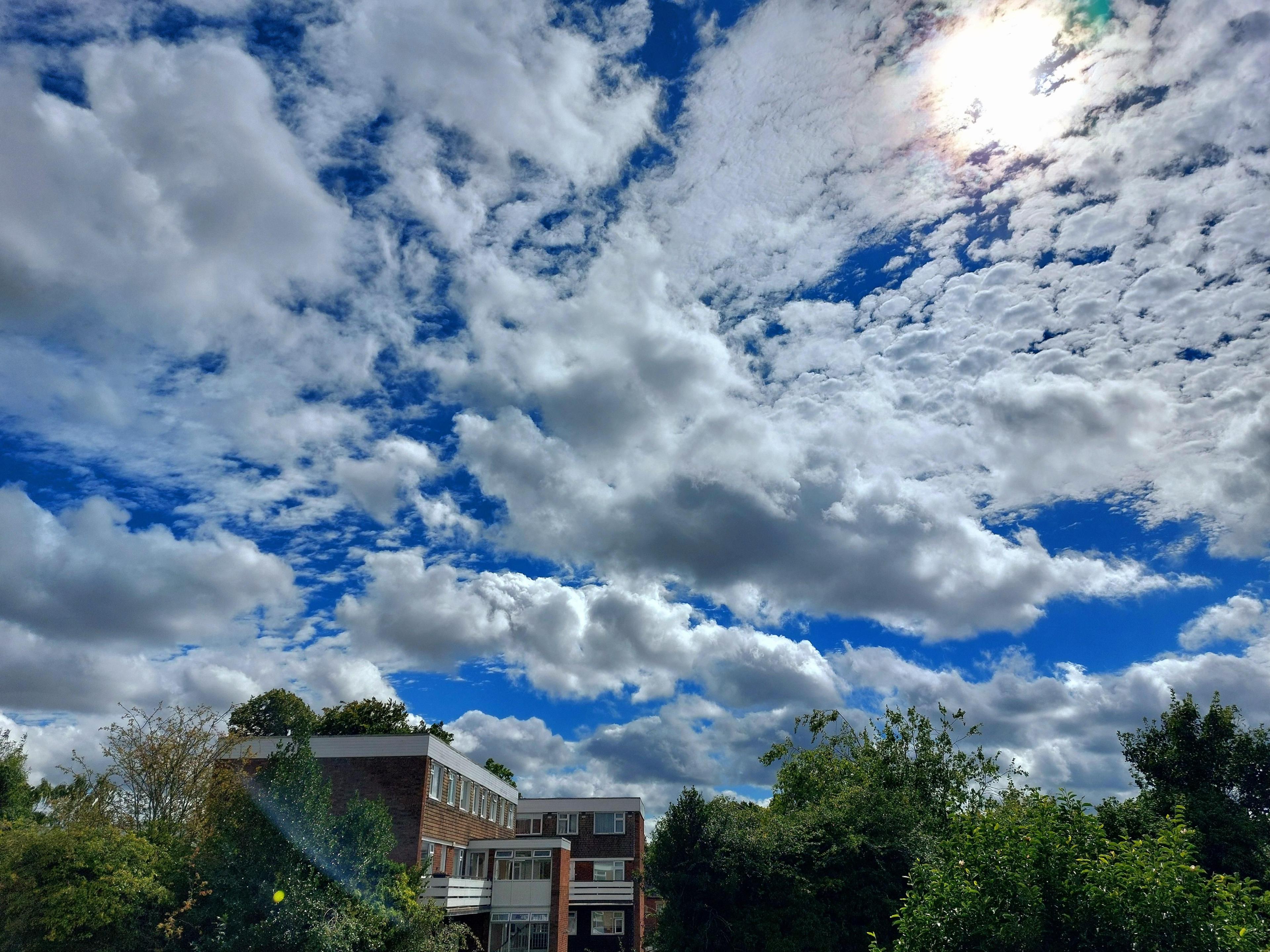 Clouds sprawled across bright blue sky with a red brick building and greenery below