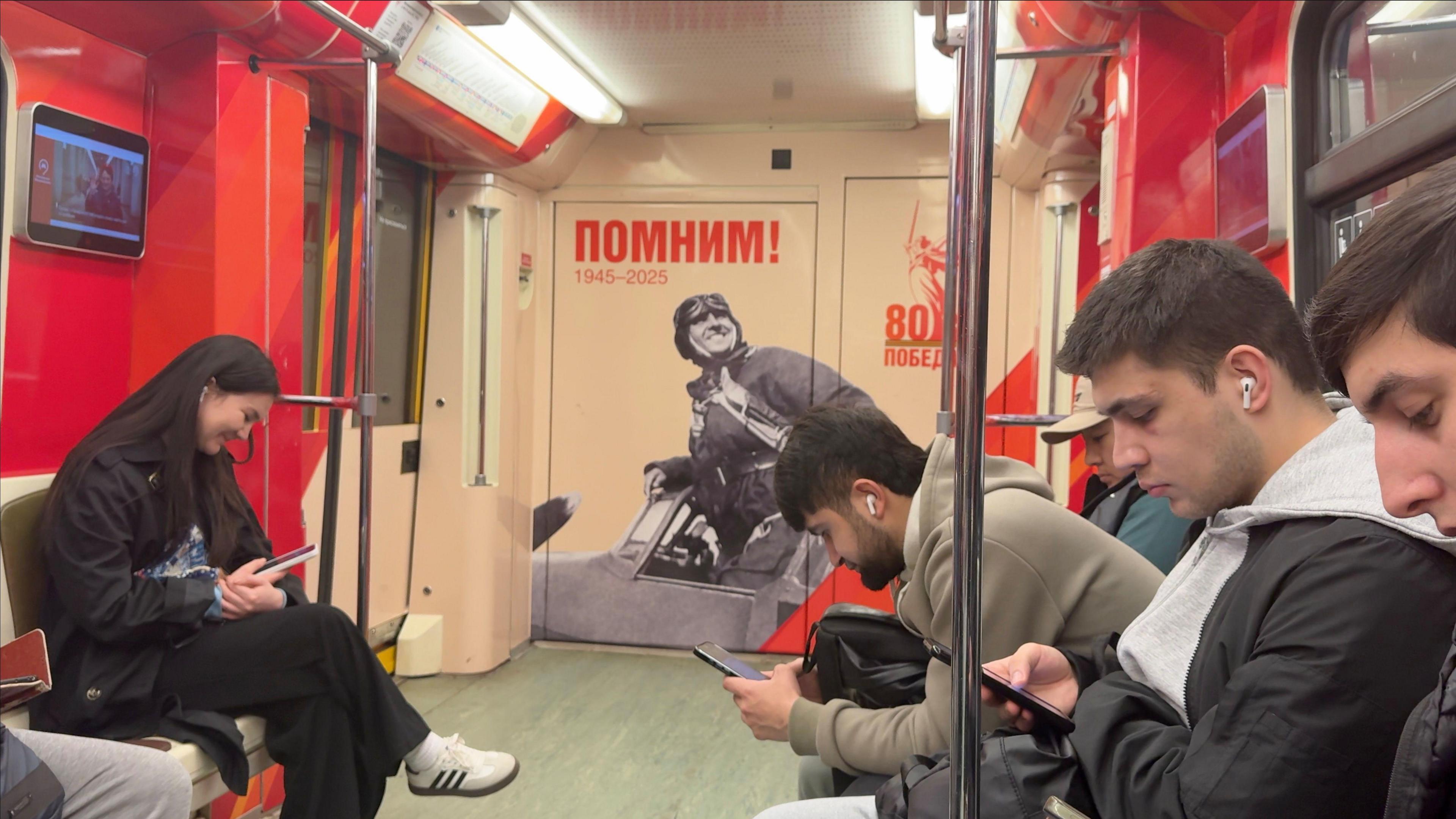 The inside of a Moscow metro carriage. The passengers are look at their phones but the back wall of the carriage has a black and white photo of a World War Two pilot and a logo celebrating the 80th anniversary of Russian victory.