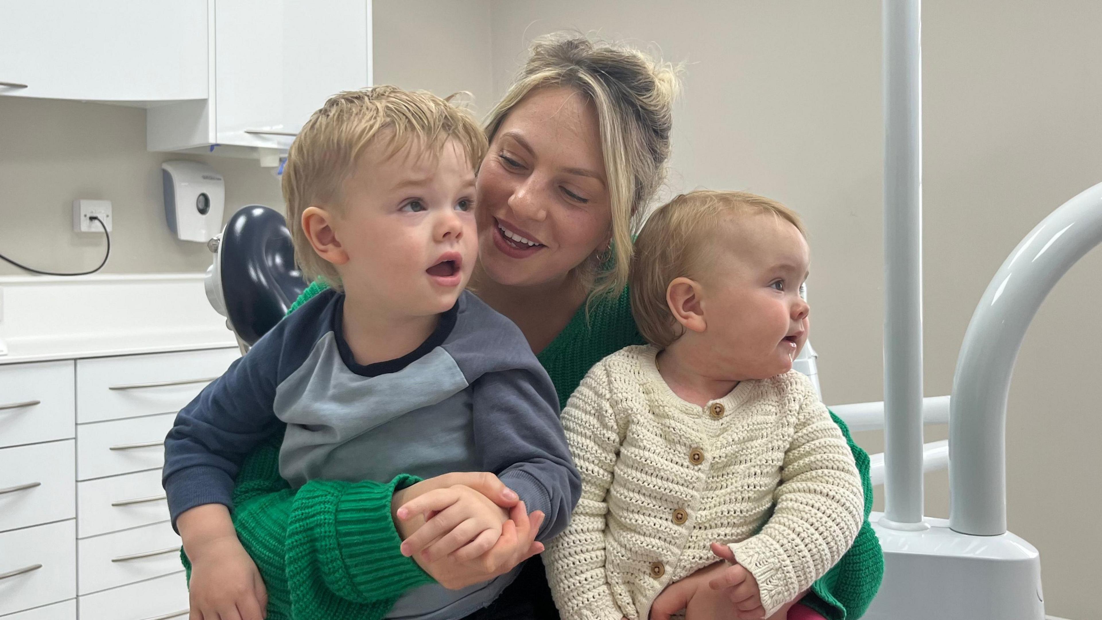 Mother smiling with her two young children on her lap, as she sits in a dental surgery