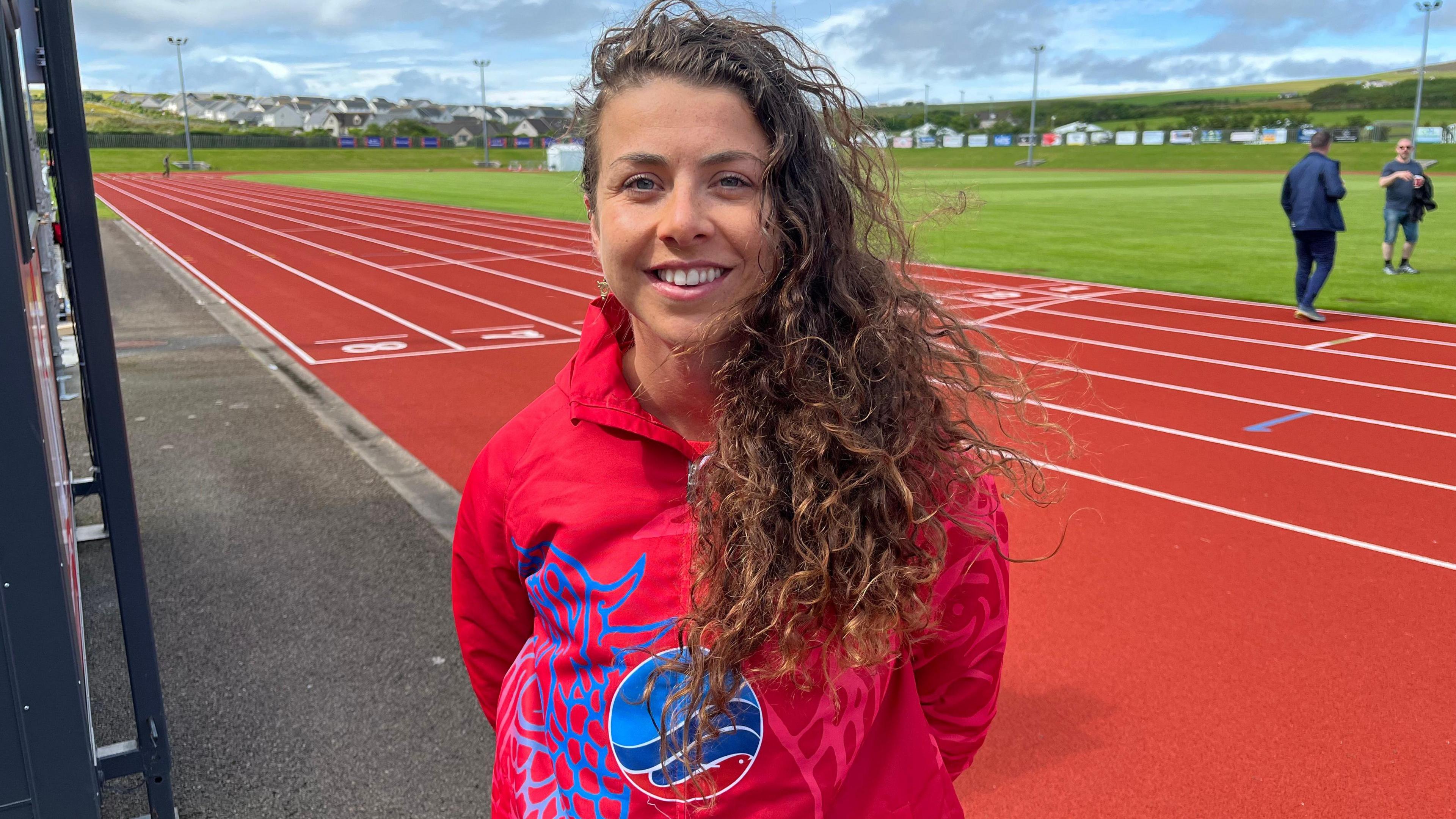 A woman with long, curly brown hair swept to one side stands in front of a red athletics track. She is wearing a red tracksuit top.