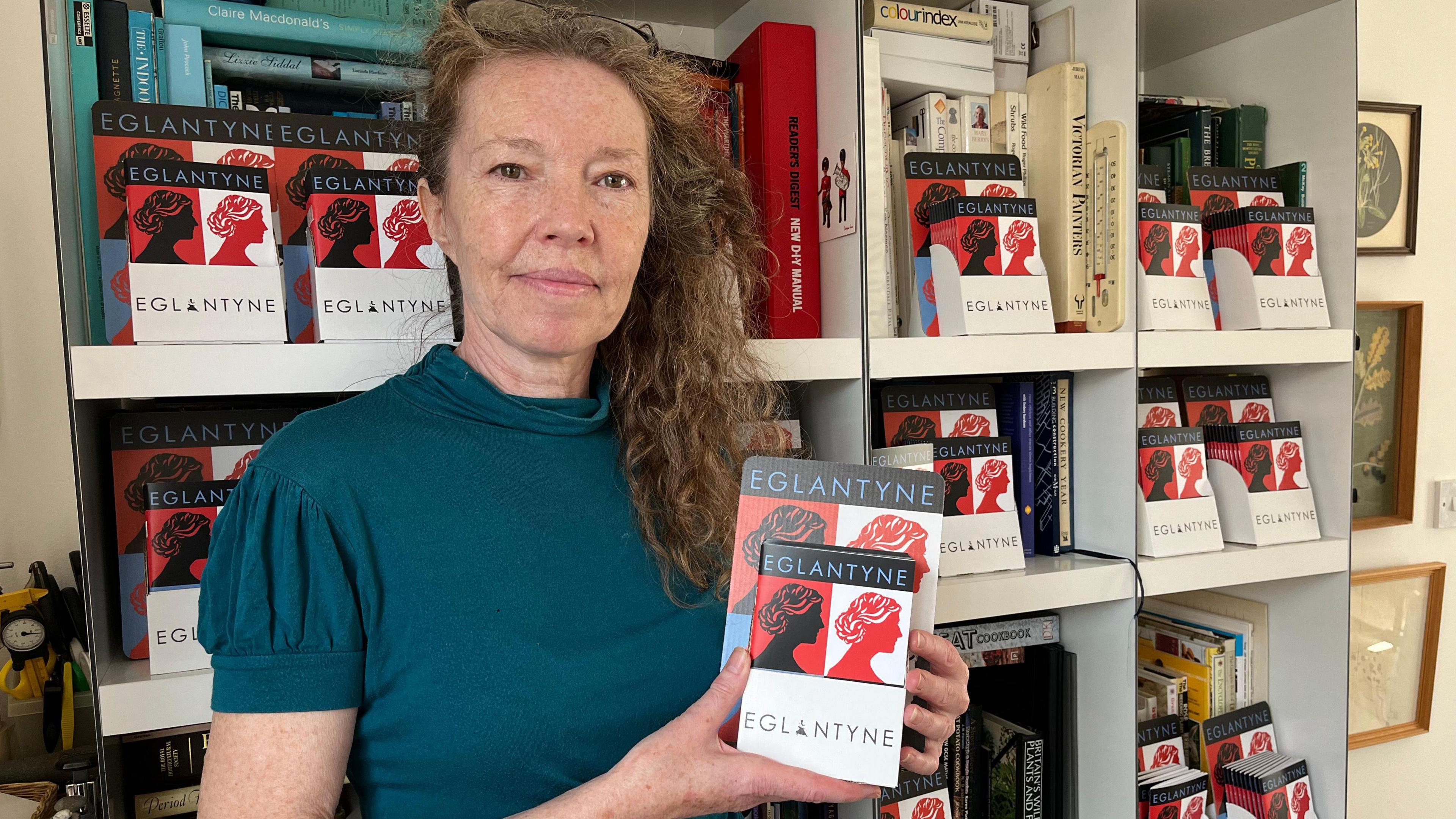 A woman with long curly hair swept to one side in a teal short-sleeved top, holding a book titled Eglantyne.