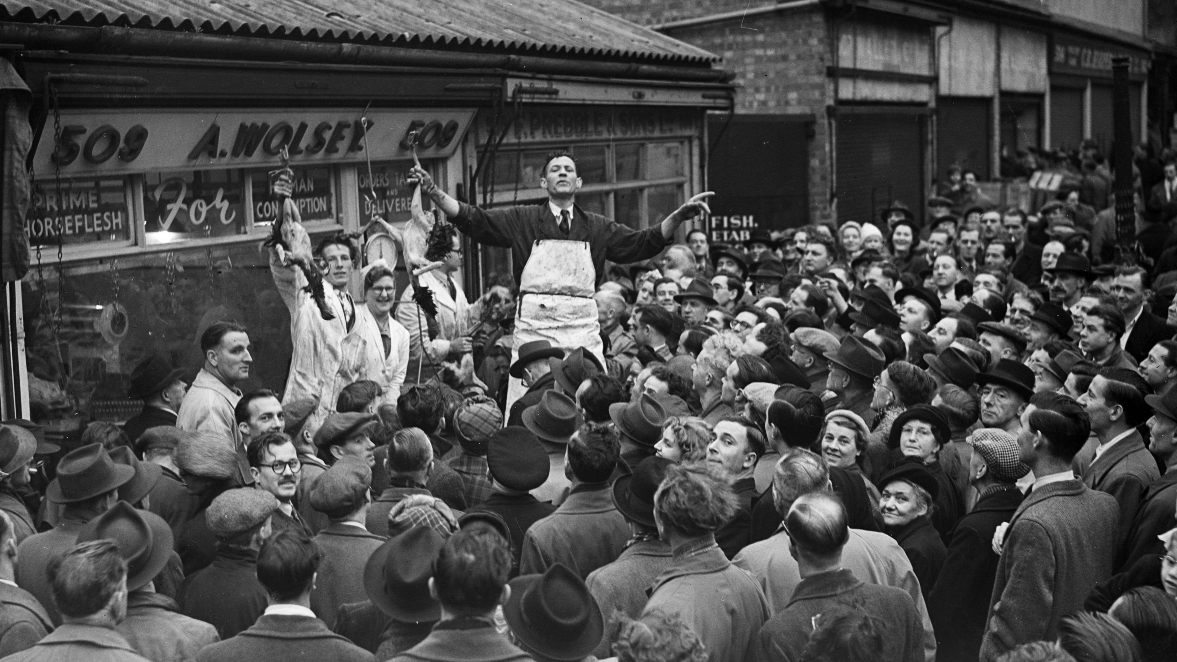 Black and white image of crowds of people surrounding four traders auctioning off poultry