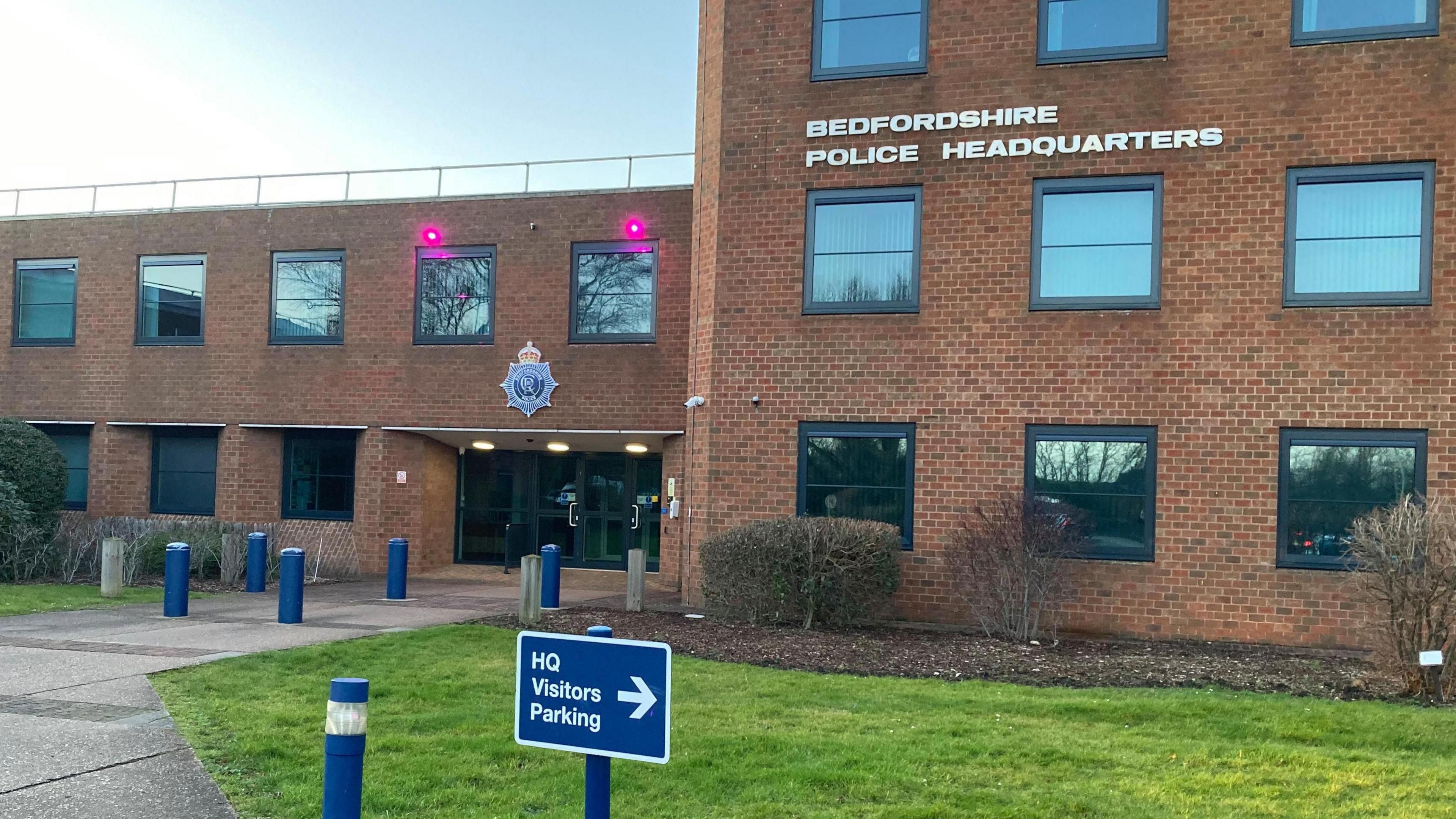 The front of Bedfordshire Police headquarters. It is a red brick building with grass and bushes outside. In the foreground there is a sign for visitors parking.