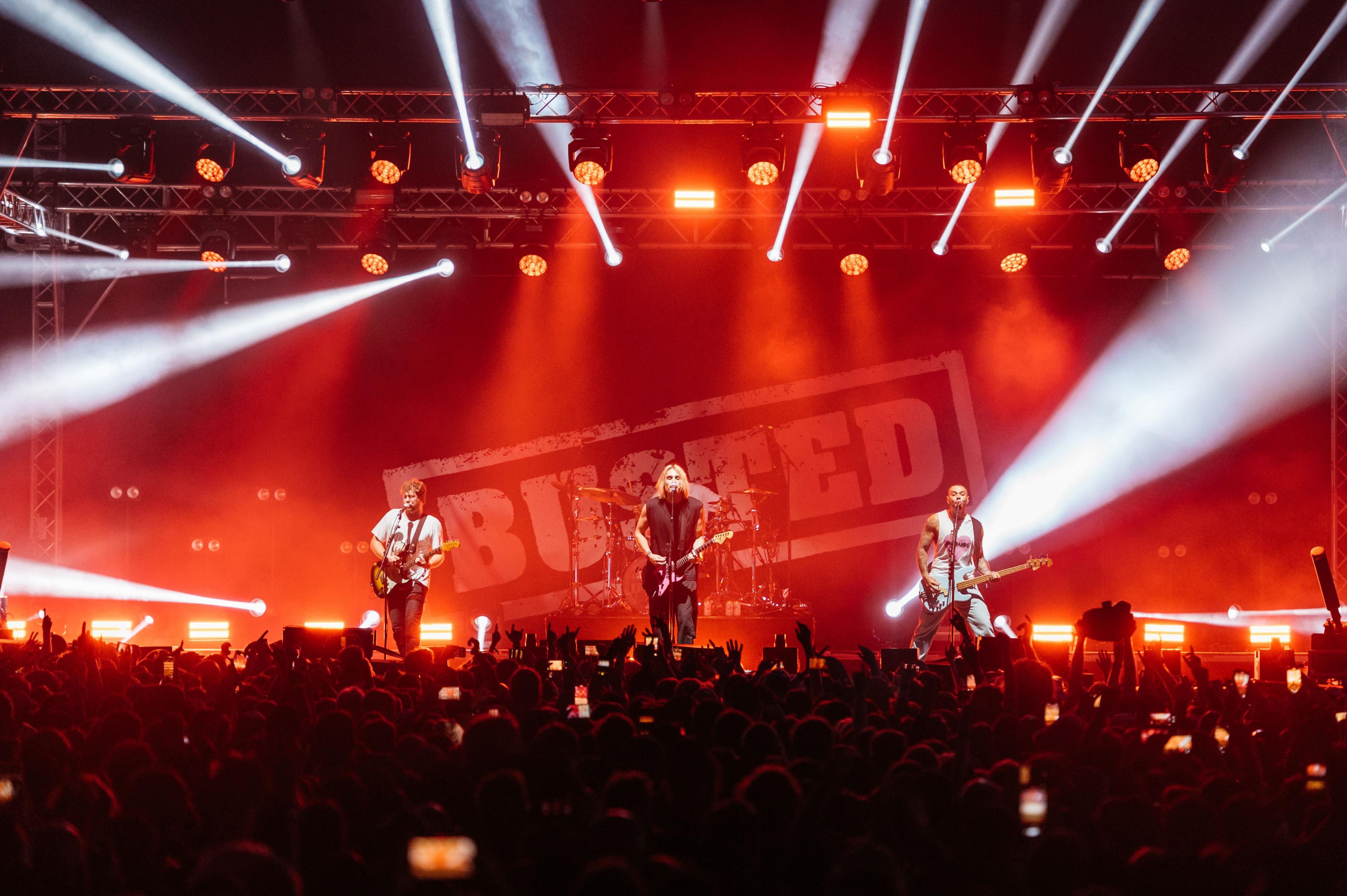 Busted performing on stage to a crowd. There are red and white lights coming from the stage and they are all singing and playing guitars.