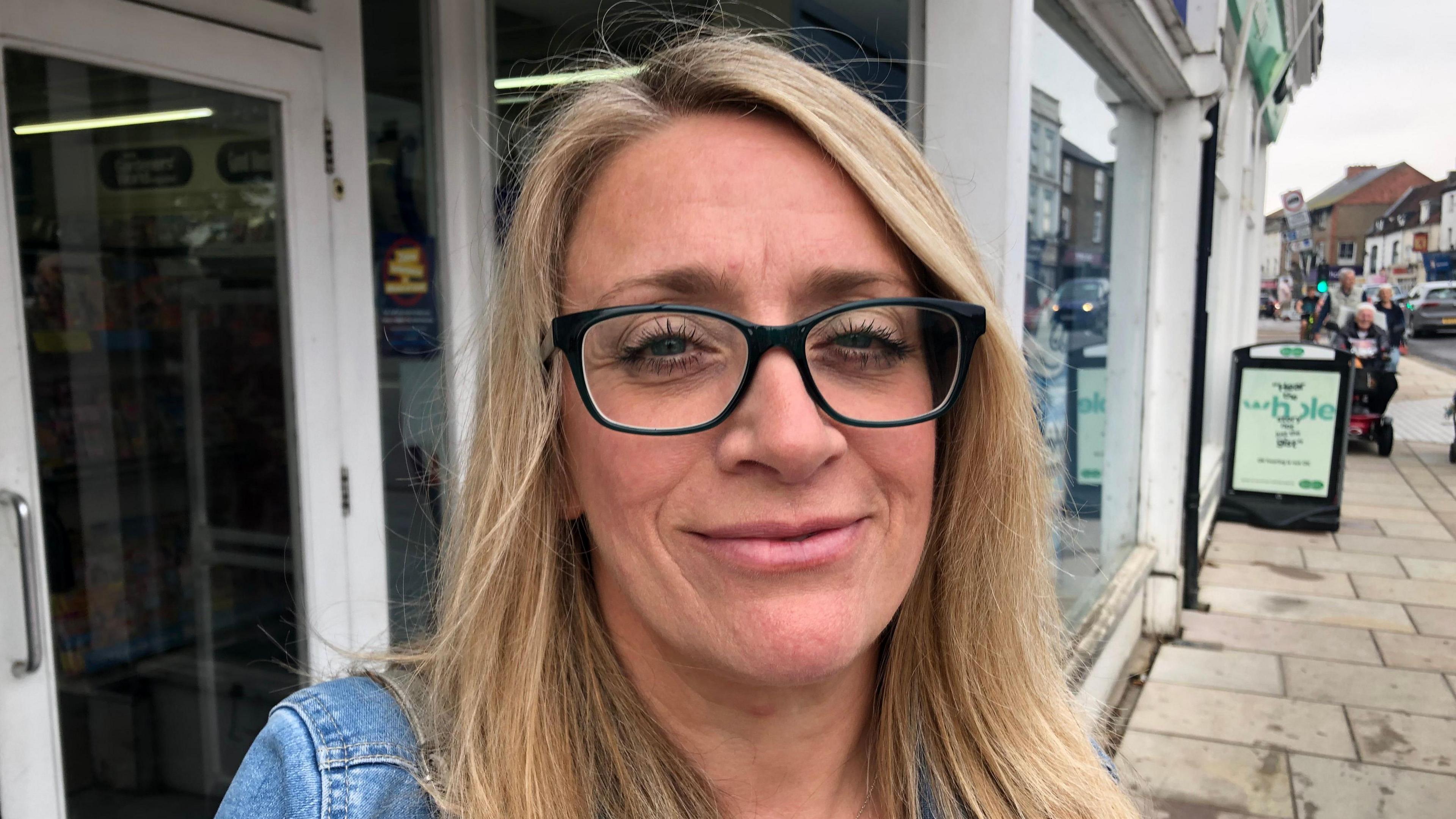 Christine Lewis is blonde and wearing glasses with black frames. She is smiling at the camera and wearing a denim jacket.