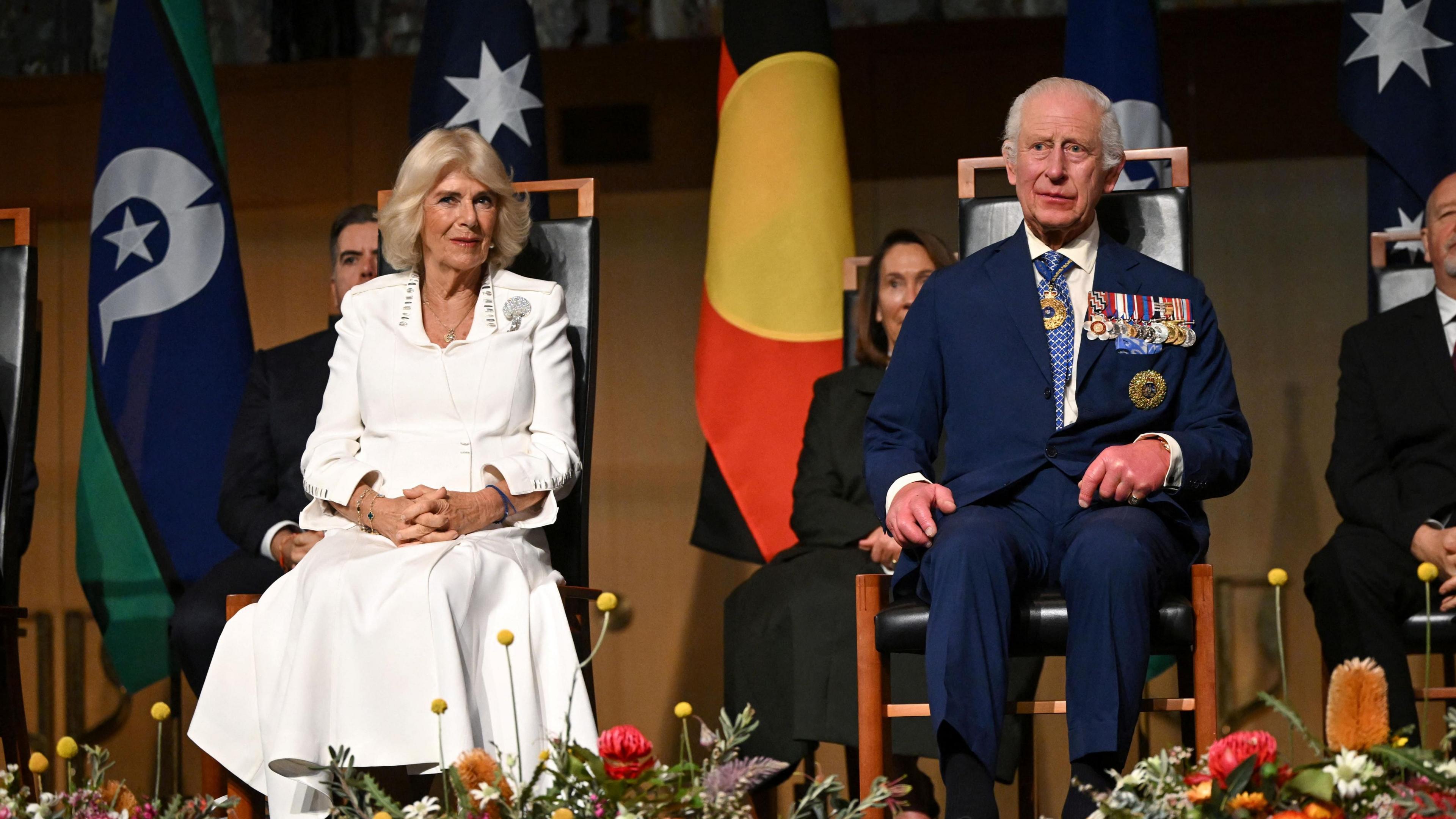 King Charles and Queen Camilla sat in wooden chairs on a stage with flowers at their feet.