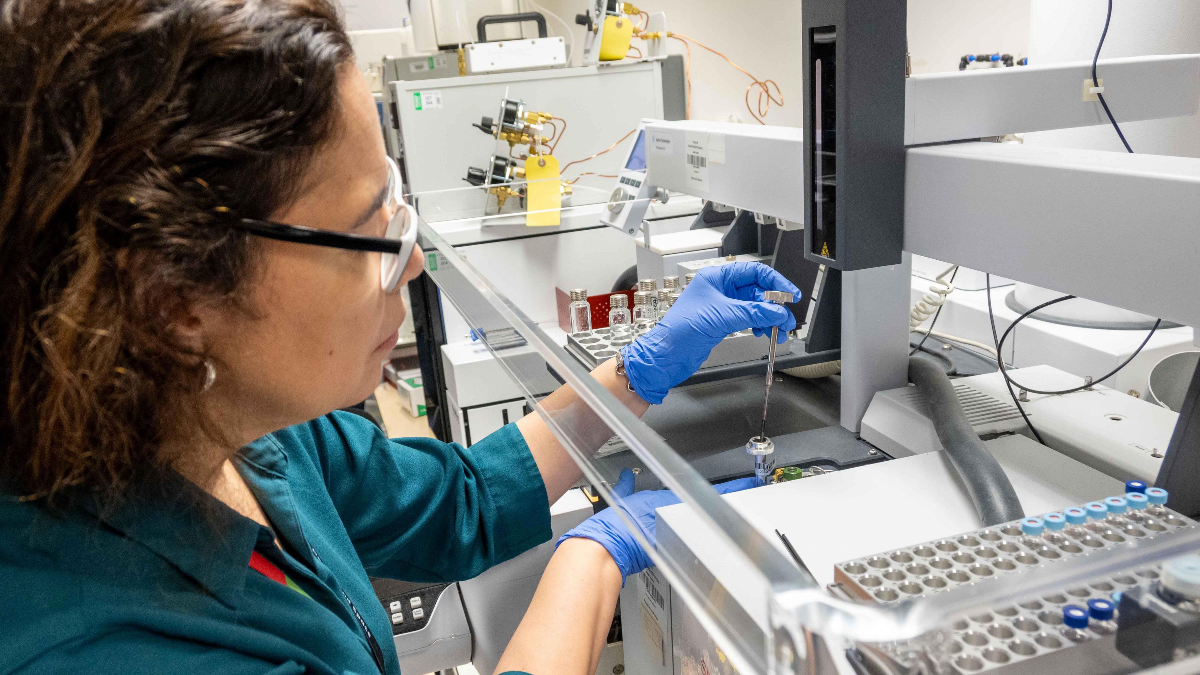 Dr Luciana da Costa Carvalho sits at a desk in a white labratory. She is using a piece of analytical equipment. Dr Carvalho has short brown hair and wears black framed glasses. She is wearing a teal blue coloured shirt and pale blue disposable gloves. Her right hand is holding a sample, while her left hand is holding a pipette over the sample.