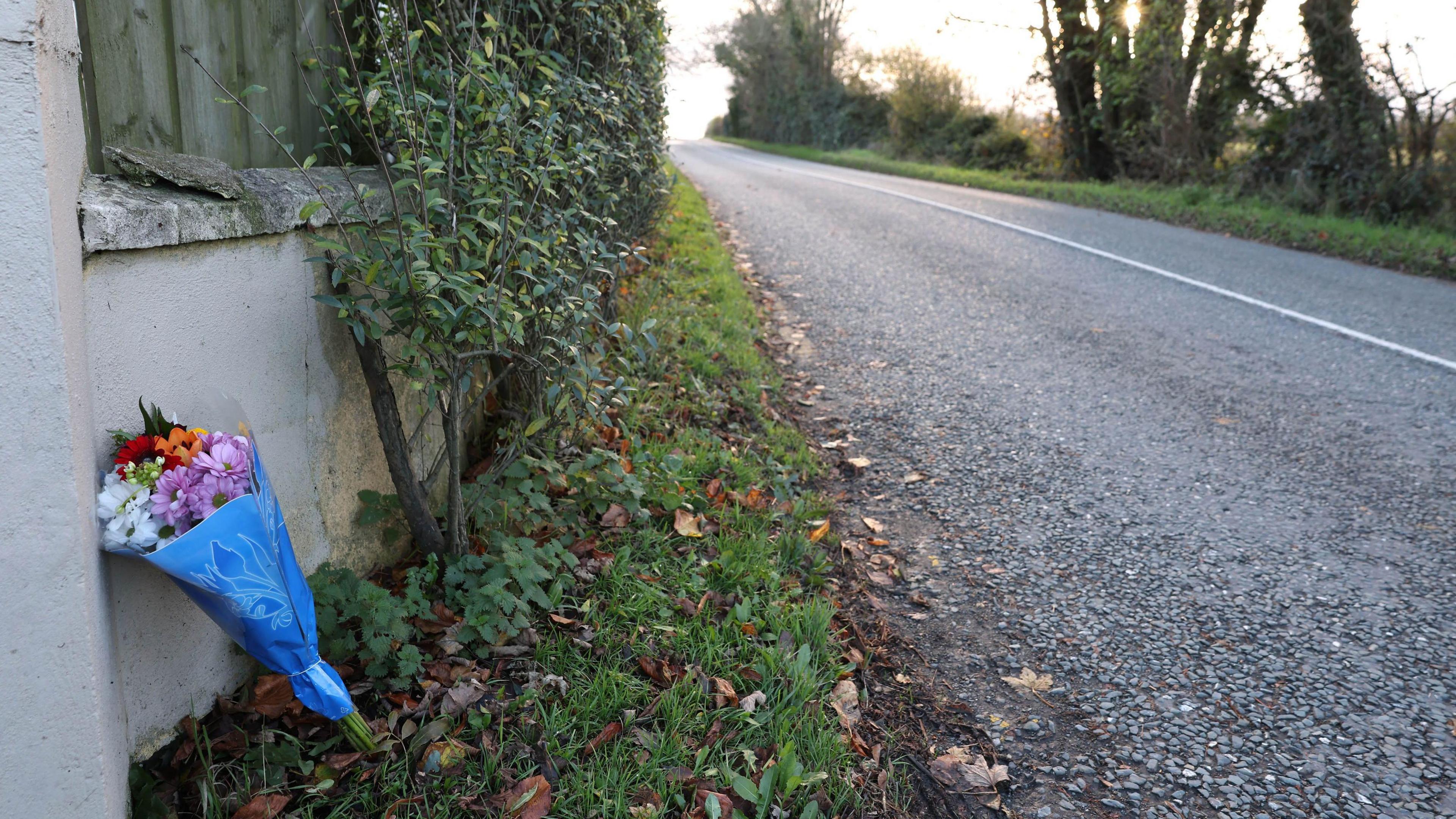 Flowers wrapped in blue wrapping placed against a wall with fencing by the side of a road.