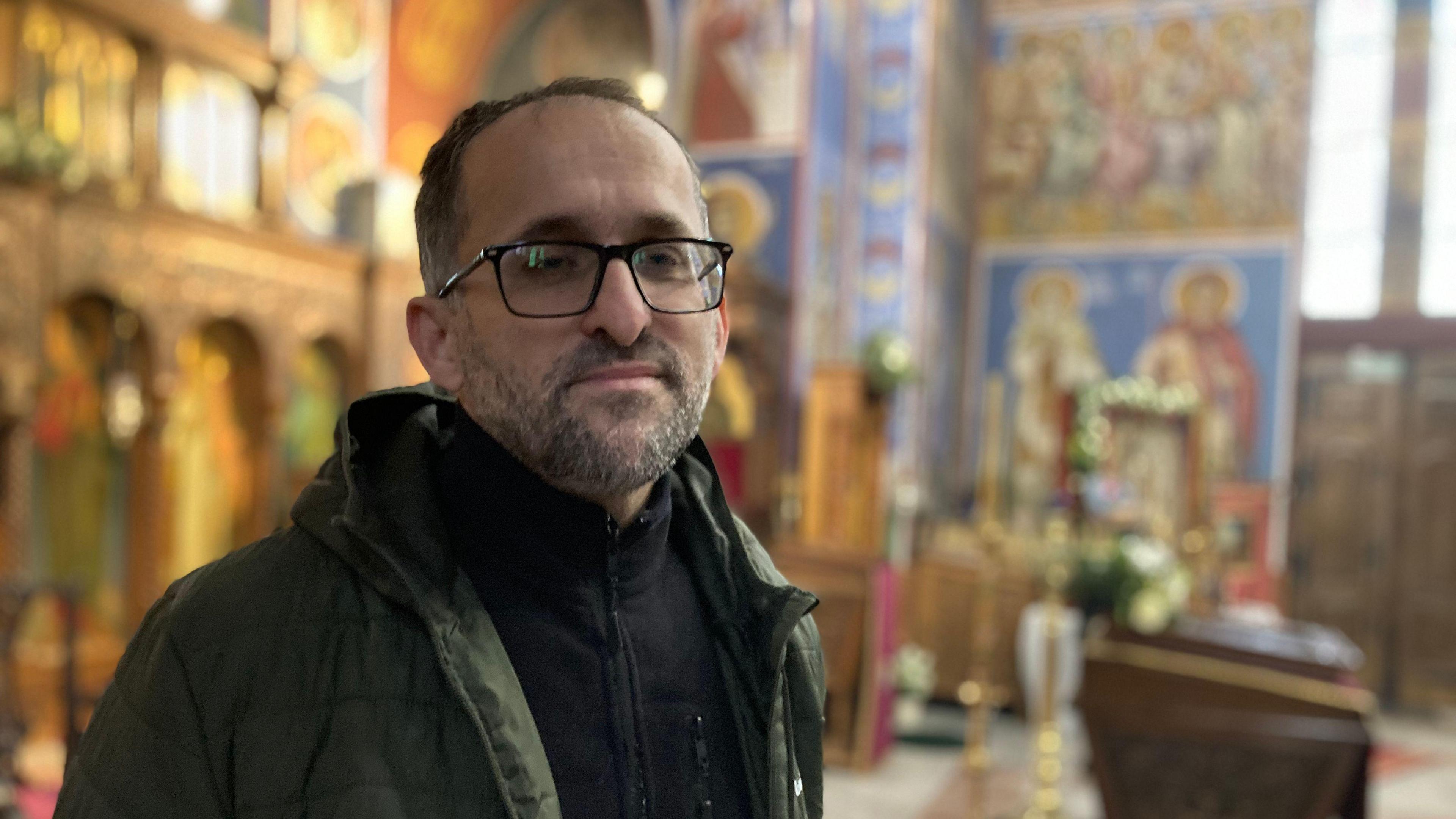 A man in a dark green coat and black fleece cardigan looks at the camera. he has dark hair and is wearing glasses and behind him are the coloured frescos of the church and some furniture. 