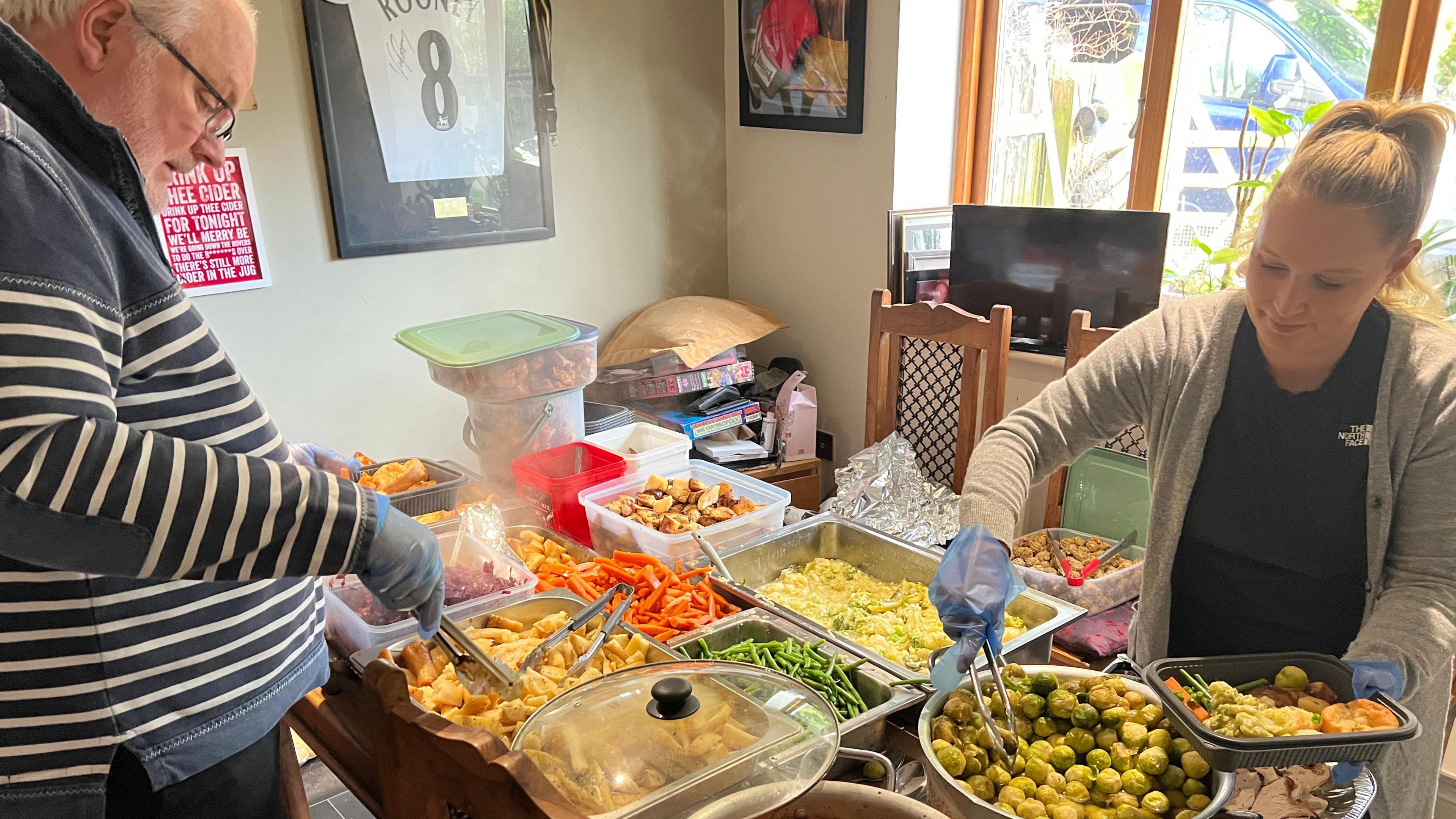 A grey-haired man in a striped jumper is holding some tongs and placing parsnips into a plastic tray. He is standing opposite a woman in a black t-shirt and grey cardigan who is serving up sprouts.