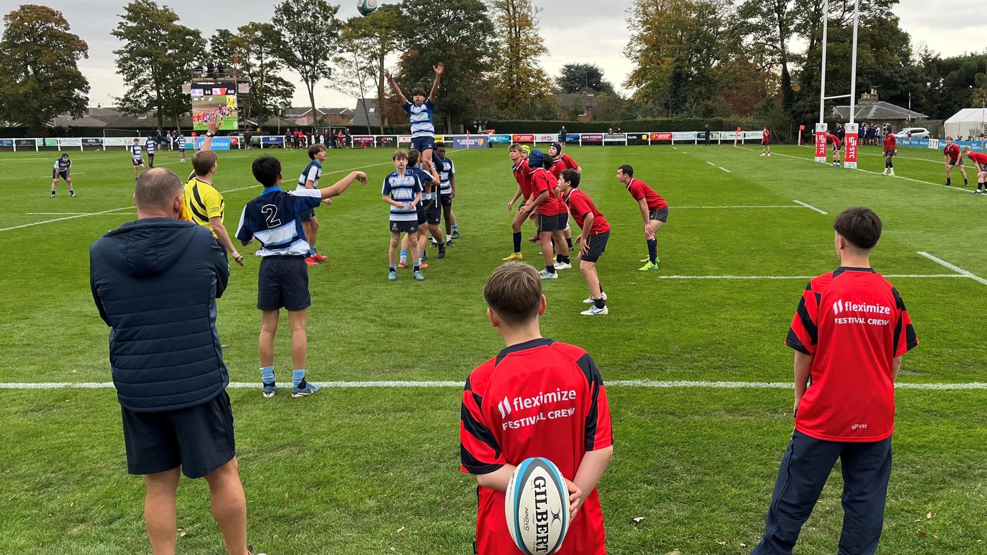 Two rugby teams were standing on a green field playing rugby. Three people stood with their backs to the camera watching them play.