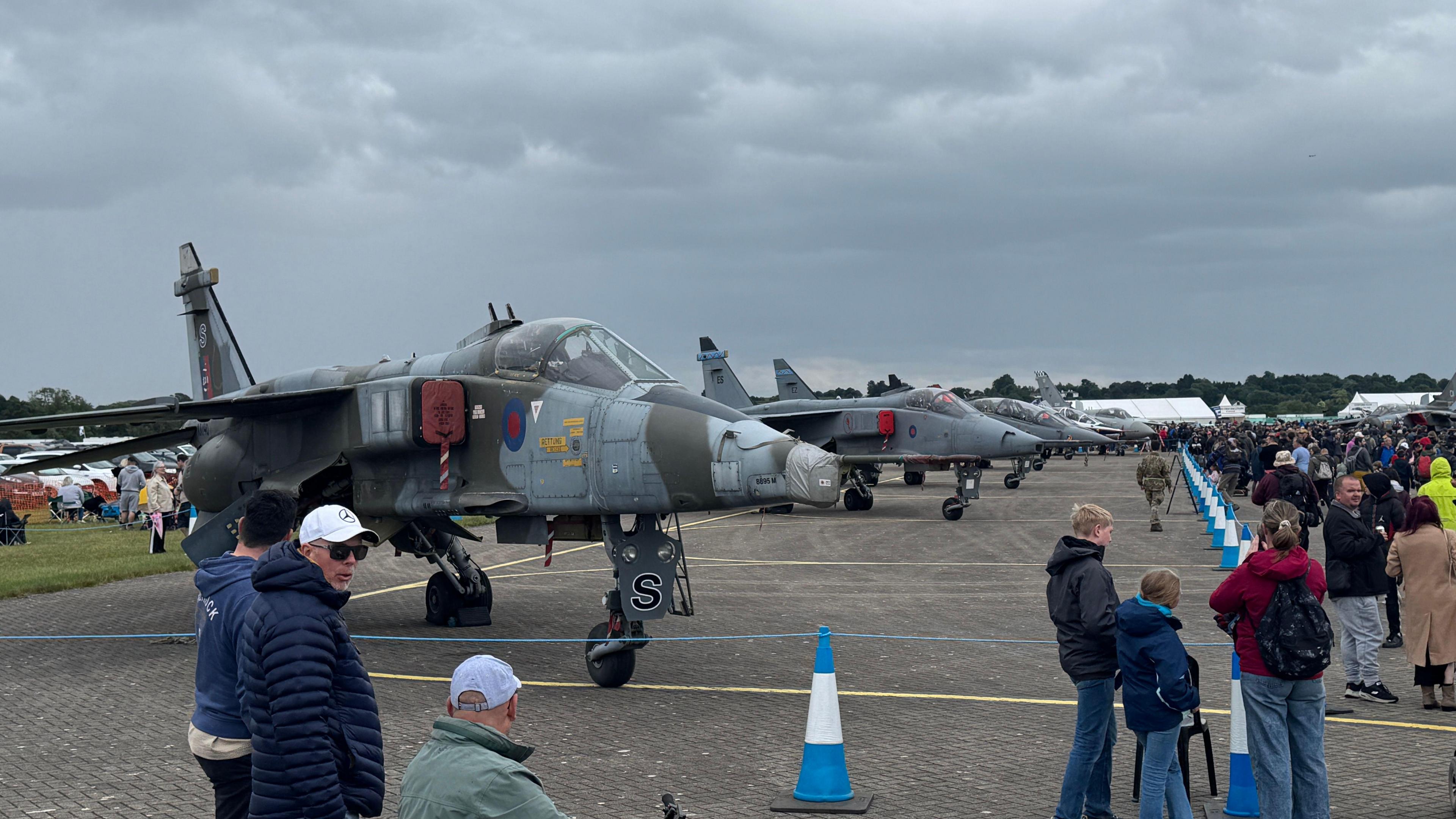 A number of fighter jets lined up in a taped off area on an airfield. People have gathered around to look at the aircraft.