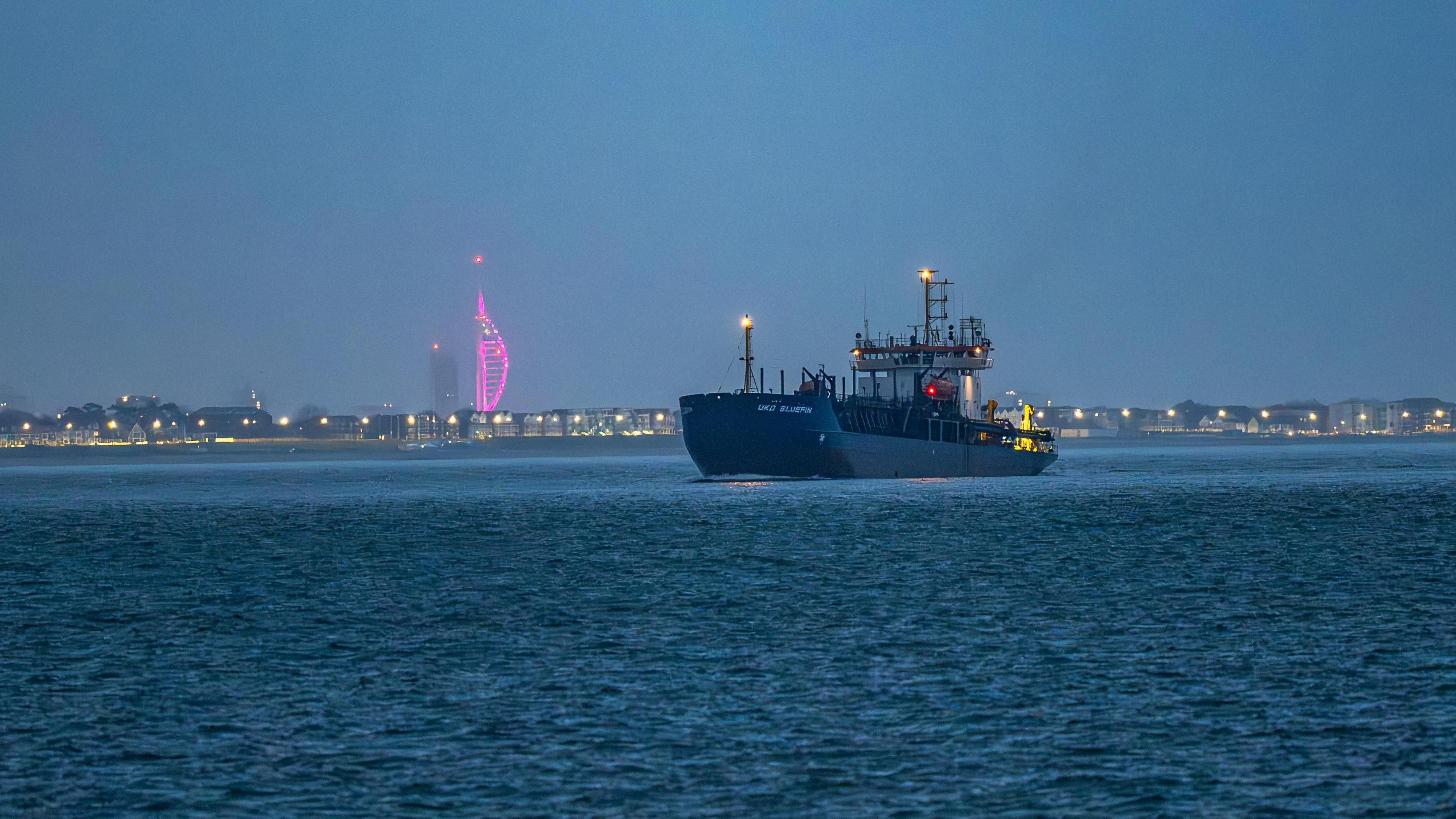 A boat on a blue sea. It is a misty morning and there are lights along the shore. In the distance the Spinnaker Tower can be seen illuminated in pink.