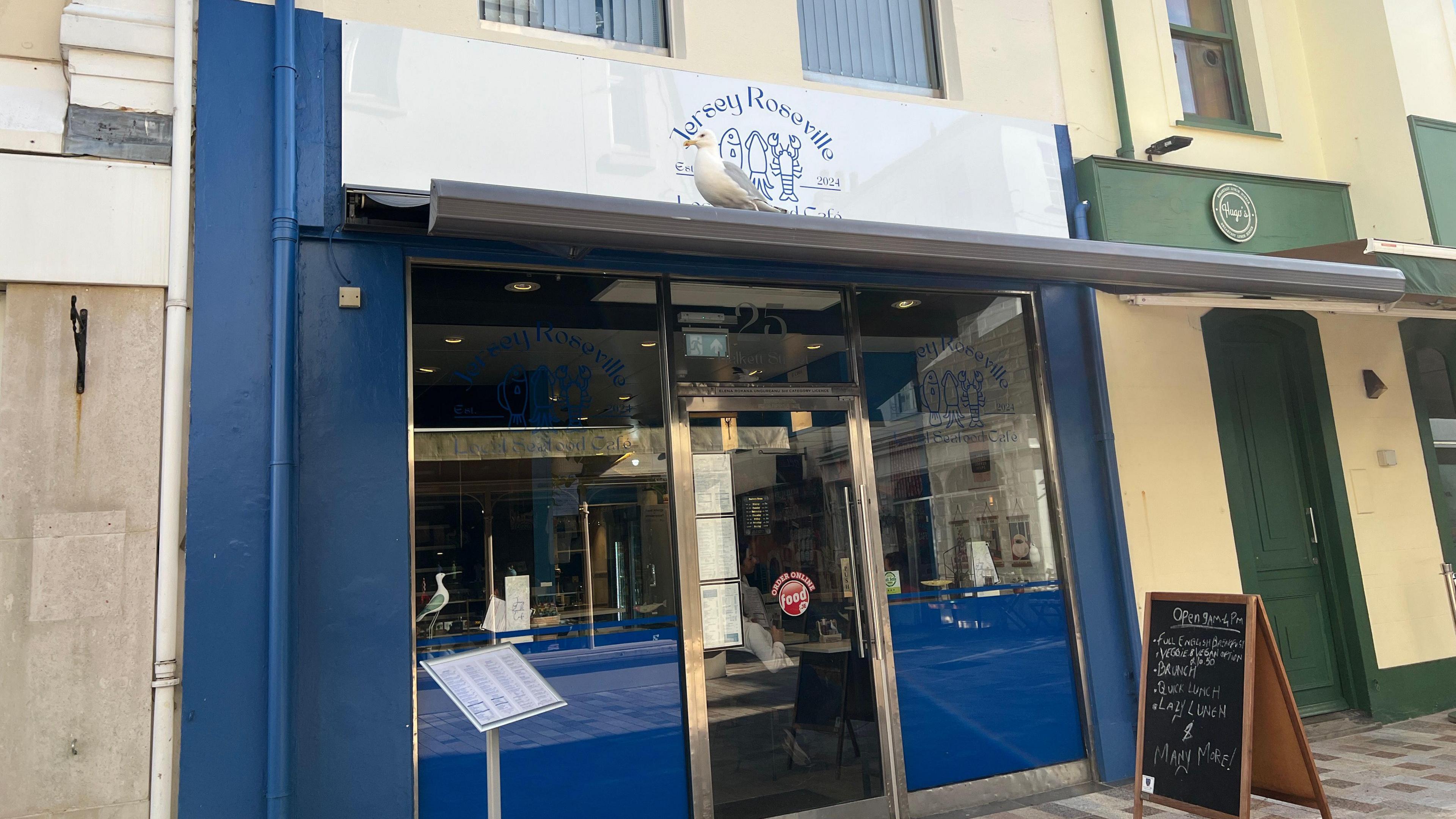 Its the outside of the cafe with which has a blackboard by the door with the menu on. The entrance is made from glass panels with white boarding over it with the cafe logo on it. There's a seagull perched on the banner over the entrance.