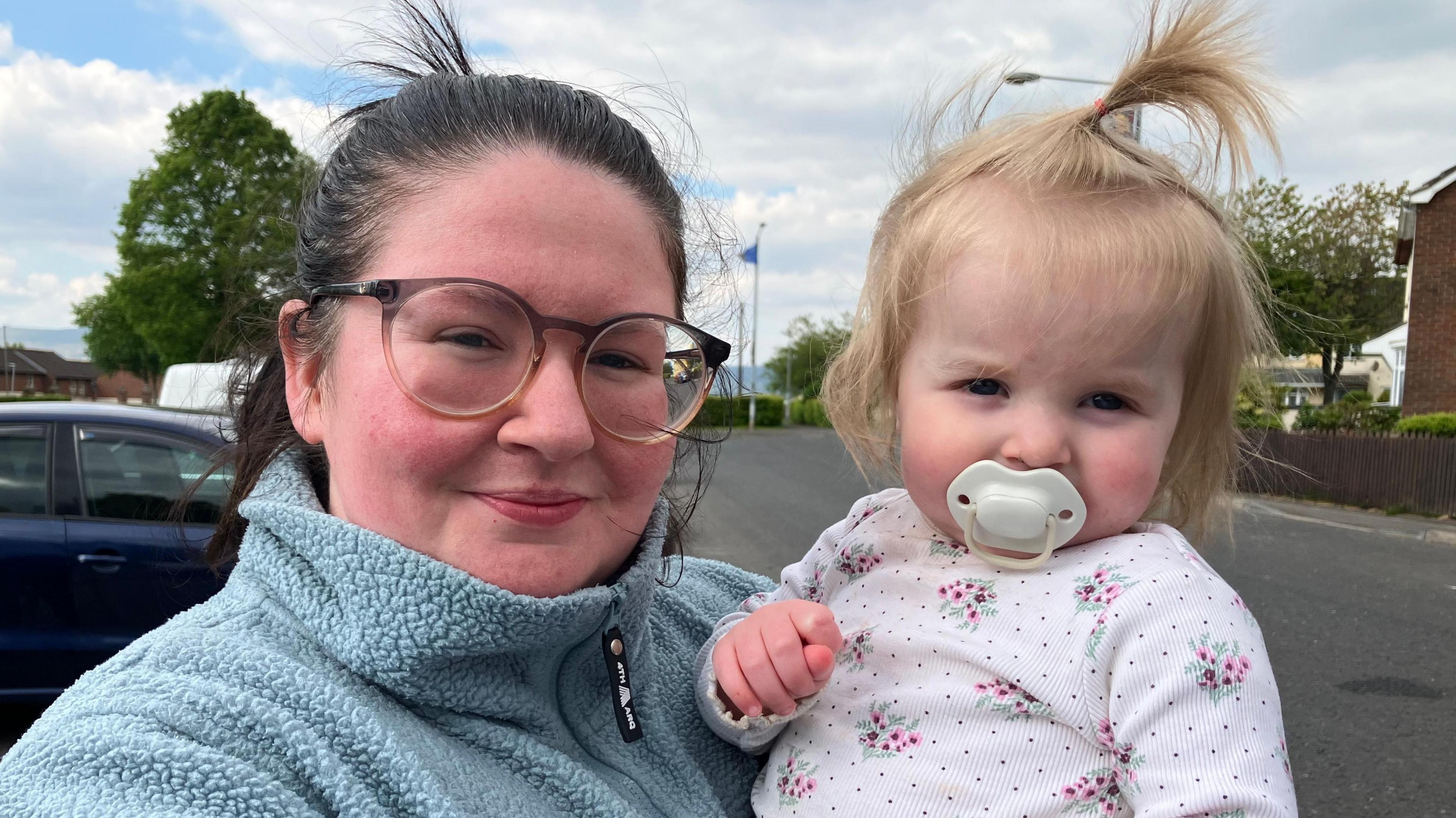 A woman with black, pulled-back hair is standing on a street, holding a small child in her arms on a street. She is wearing black-rimmed glasses and a light blue fleece. The young girl is wearing a white floral top and has a white dummy in her mouth. Her blonde hair is also tied up.