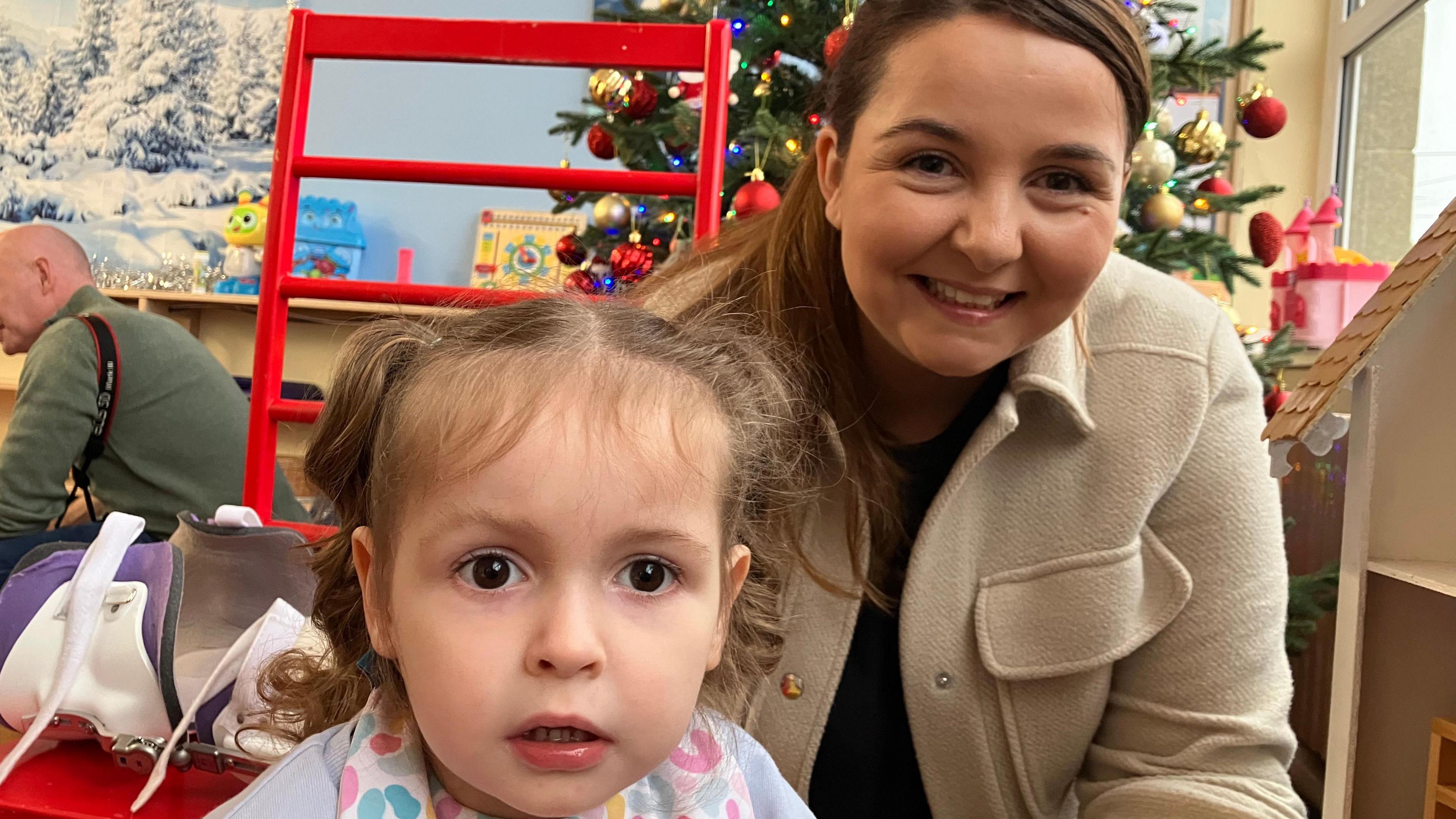 A woman with tied up brown hair is crouched down behind a child, smiling at the camera. She is wearing a dark top and a light brown jacket. Behind her is a Christmas tree. The child has fair hair, tied into pig tails, and is wearing a blue top and multi-coloured bib.  