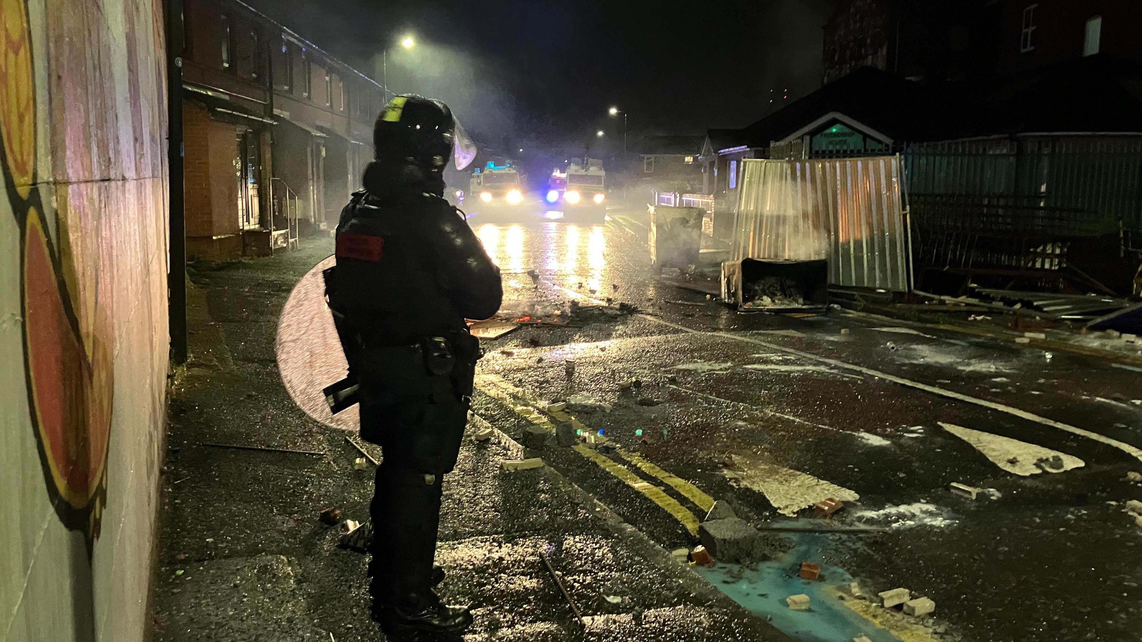 Police officer in riot gear standing looking at a street covered in debris, the lights of police landrovers in the background