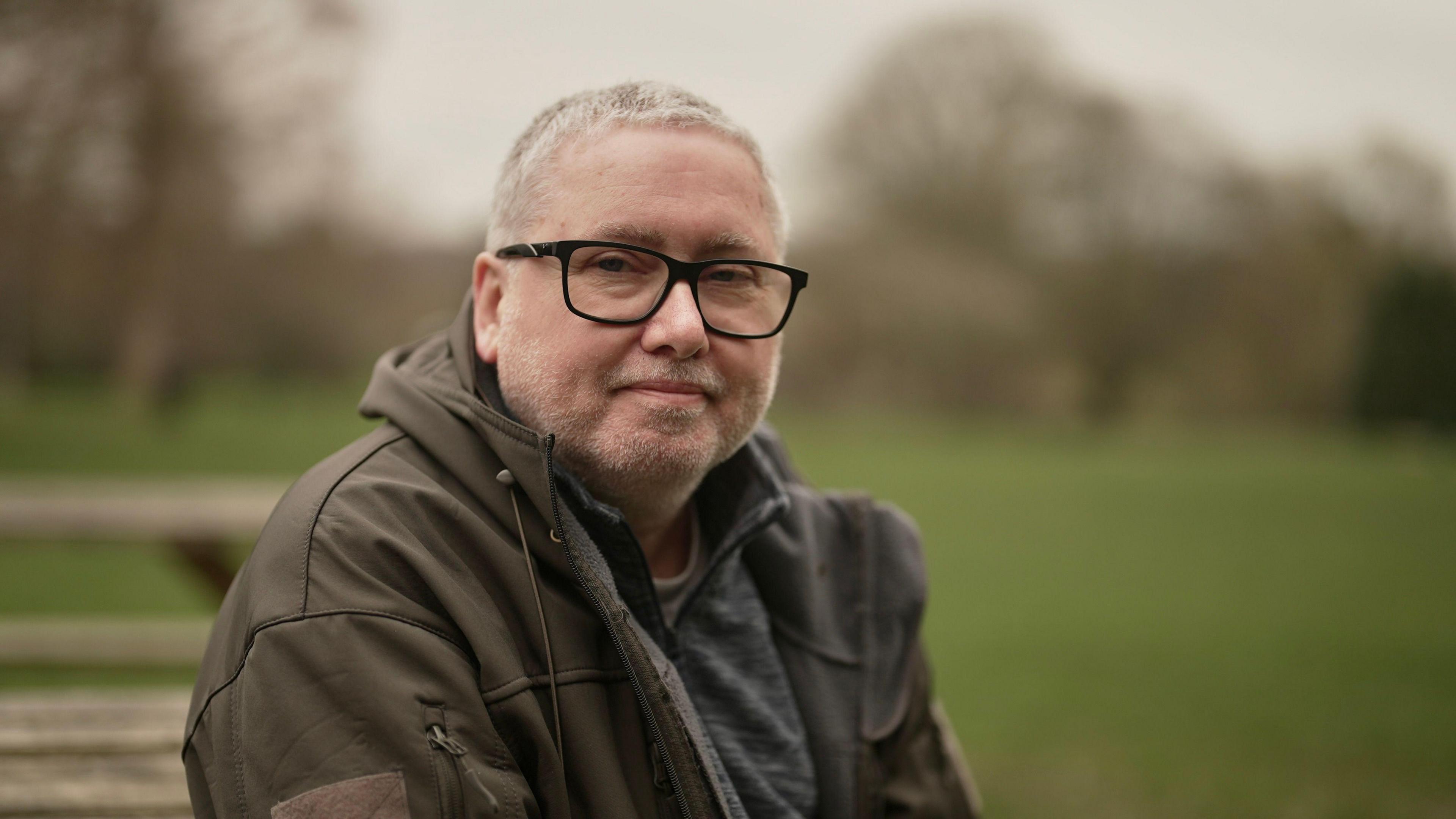 The subject is sitting down, wears glasses and a brownish/green hooded jacket.