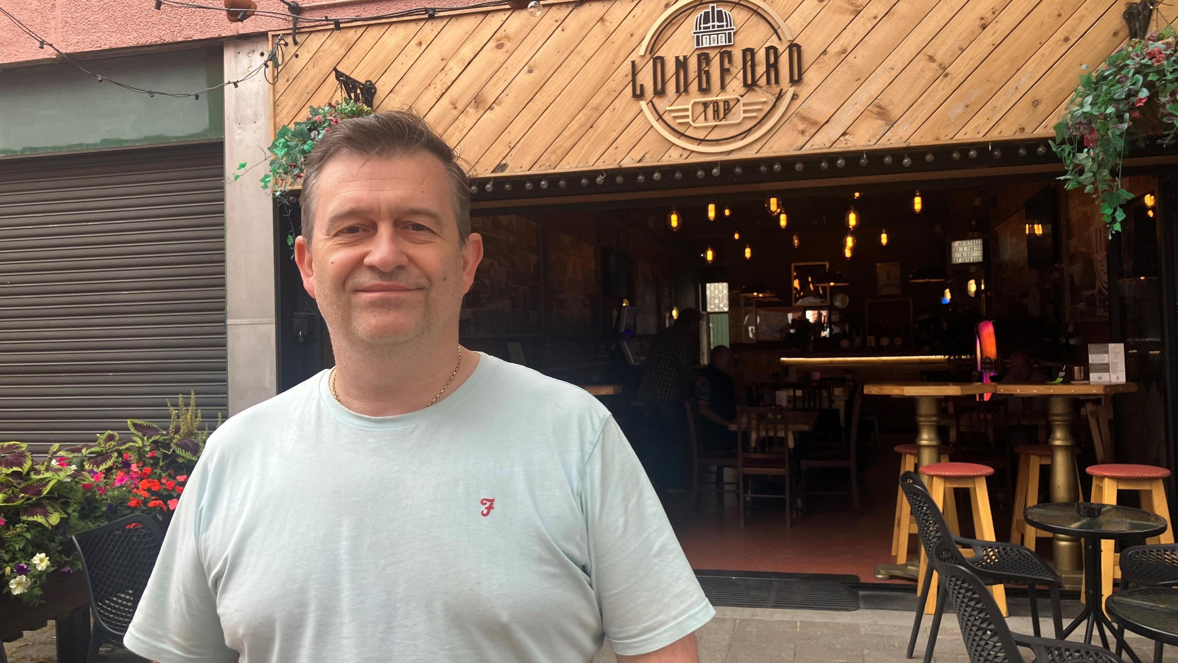 A man wearing a grey t-shirt stands smiling outside on the street in front of a pub.