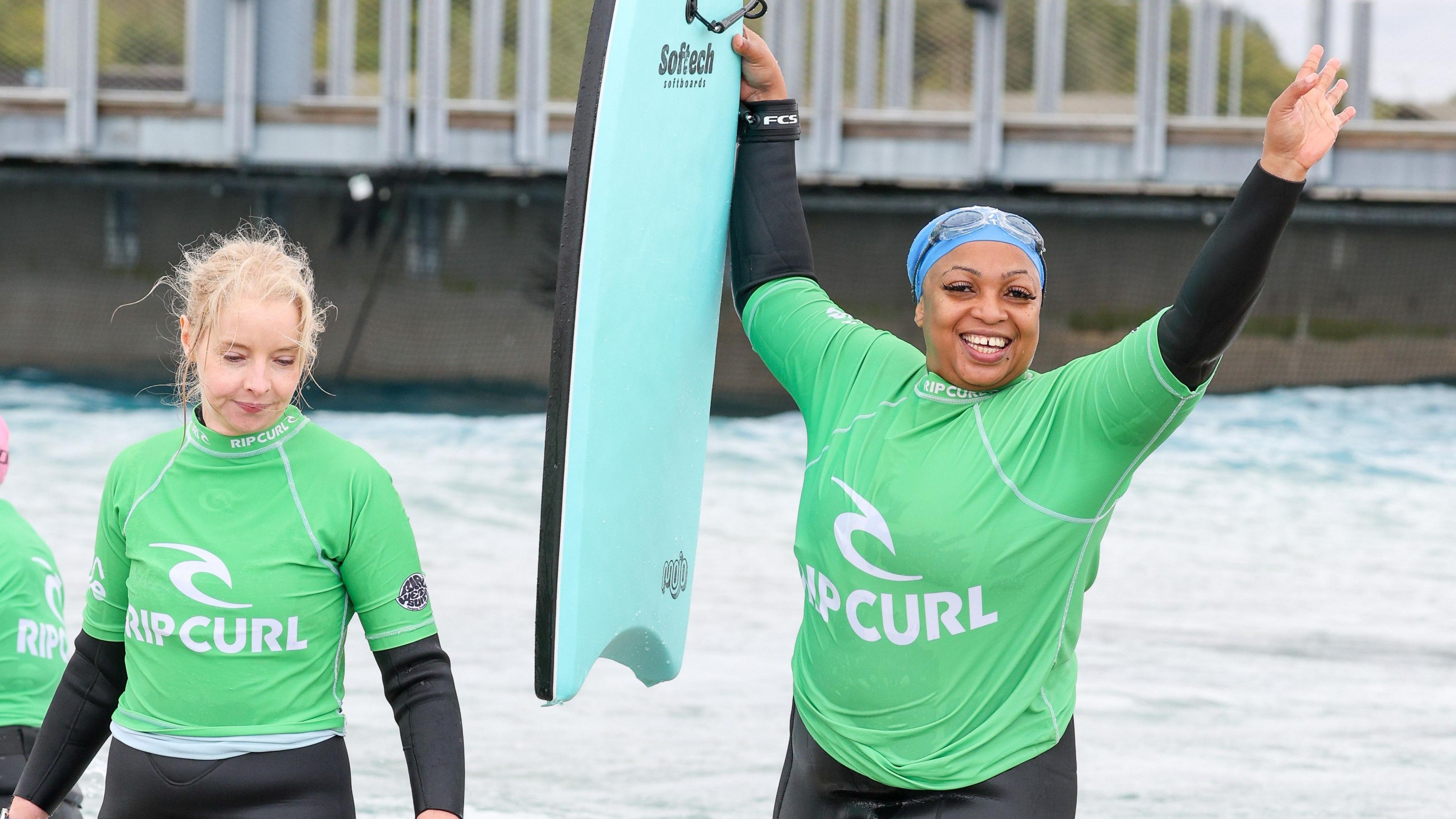 A participant waving her arms in the air and laughing. She is wearing a wetsuit, a green rash vest and a blue swimming cap with goggles. She is holding her blue bodyboard up and looks happy that she managed to catch a wave. Beside her there is another woman with her hair tied back in a bun, wearing a wetsuit and green rash vest.