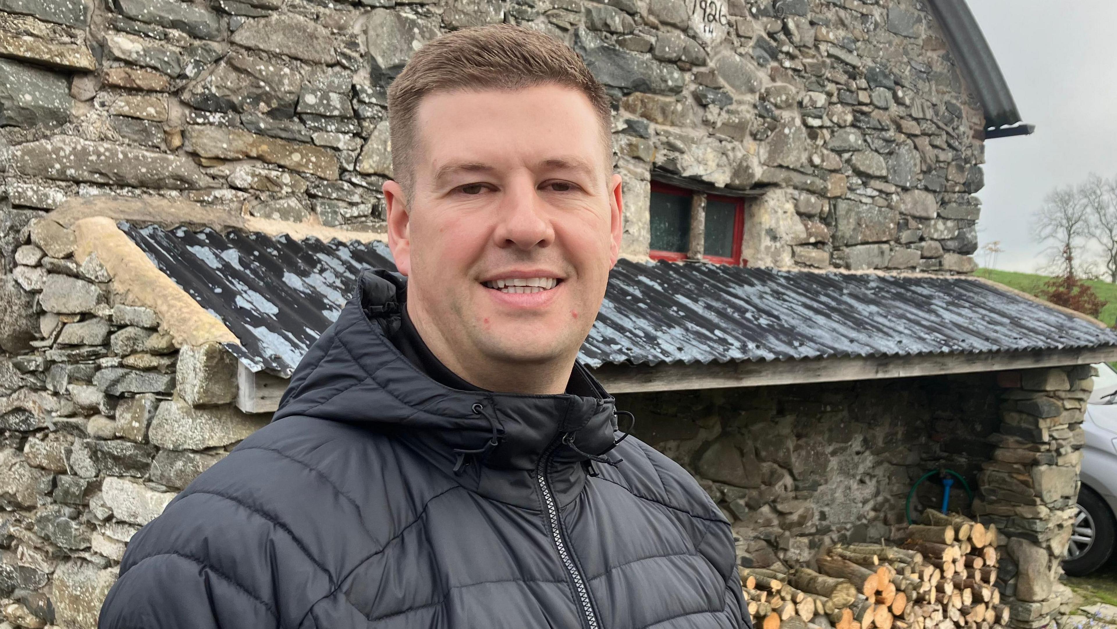 A photo of Jonny Walker who has short brown hair, and is wearing a black puffer coat standing outside in front of wood blocks on the side of a farm building. 