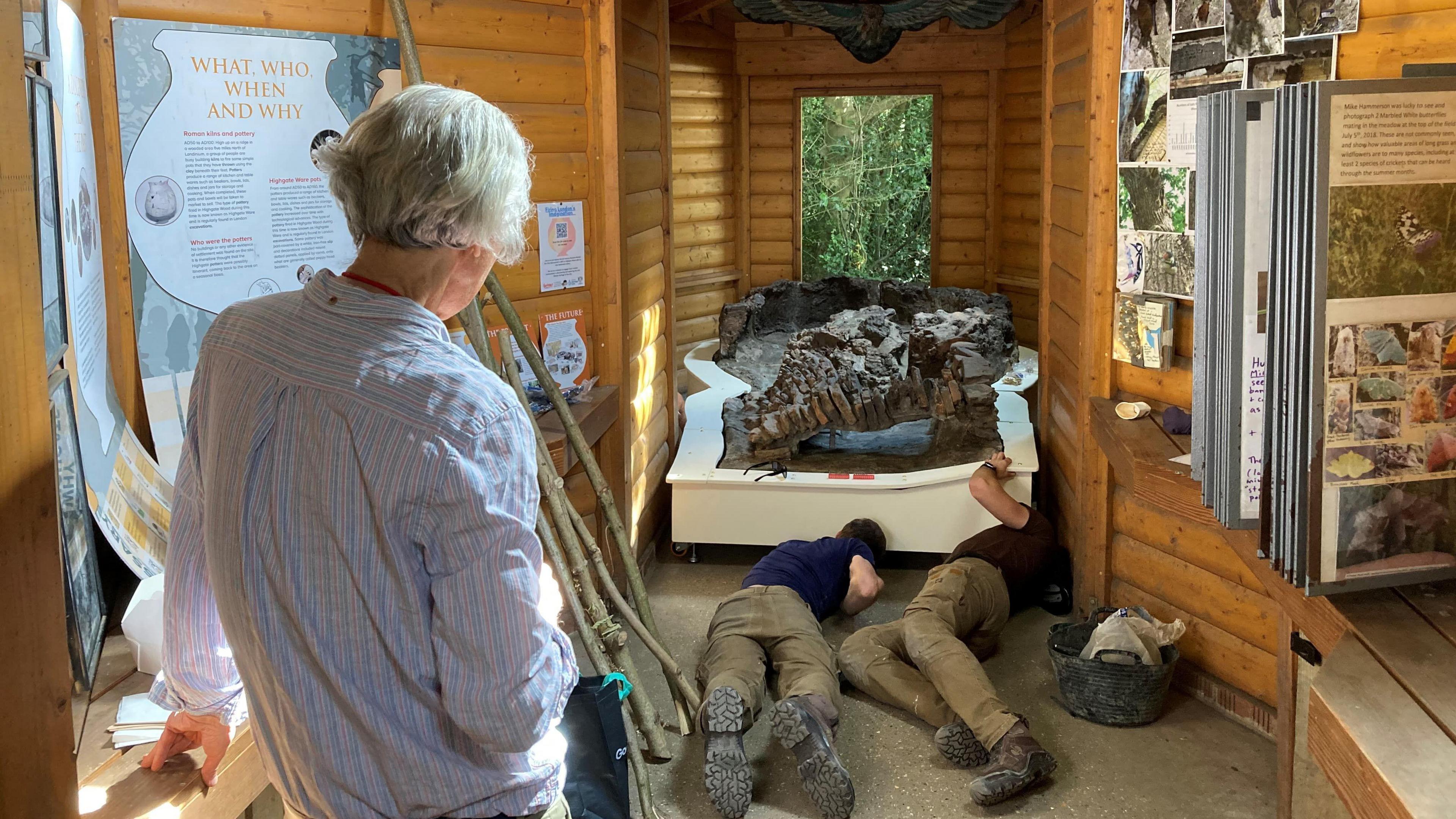 The image shows a small wooden exhibition room where two workers are on the floor, installing the ancient Roman kiln display. A person with grey hair stands nearby, watching the process. 