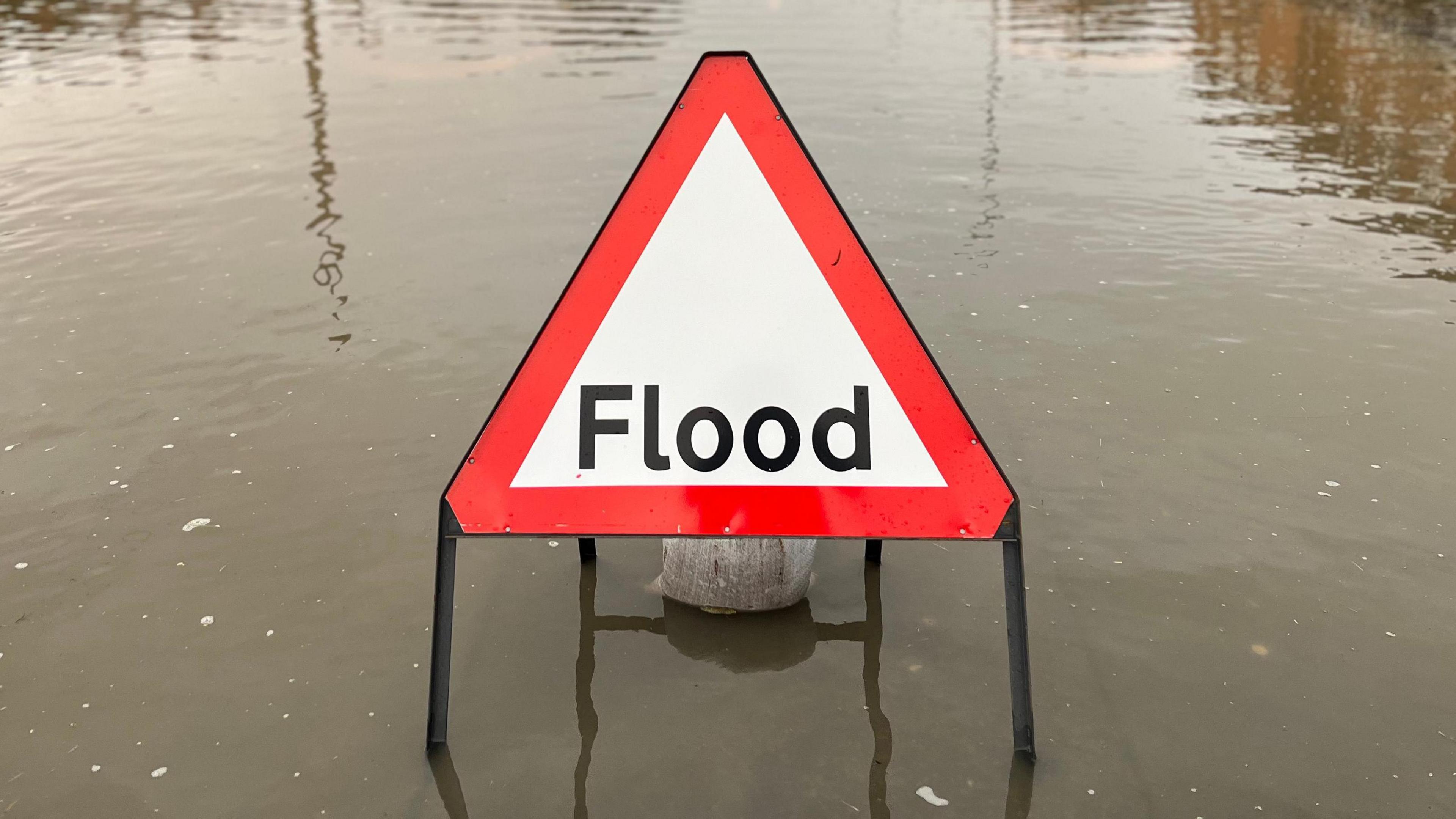A red and white flood warning sign on a flooded road. The word flood is written in black letters.