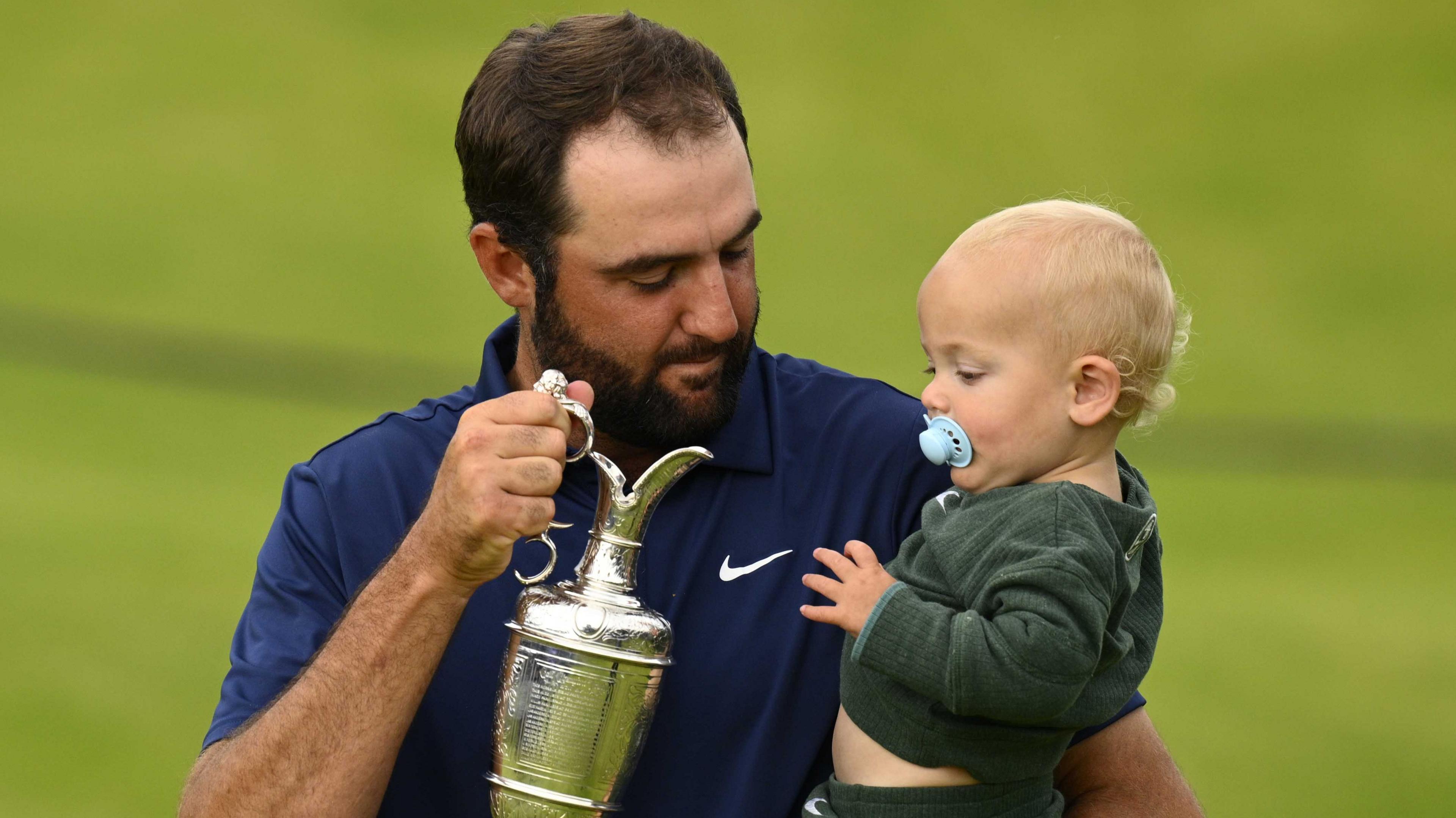 Scottie Scheffler holds the Claret Jug trophy in one hand and his baby son in the other hand.