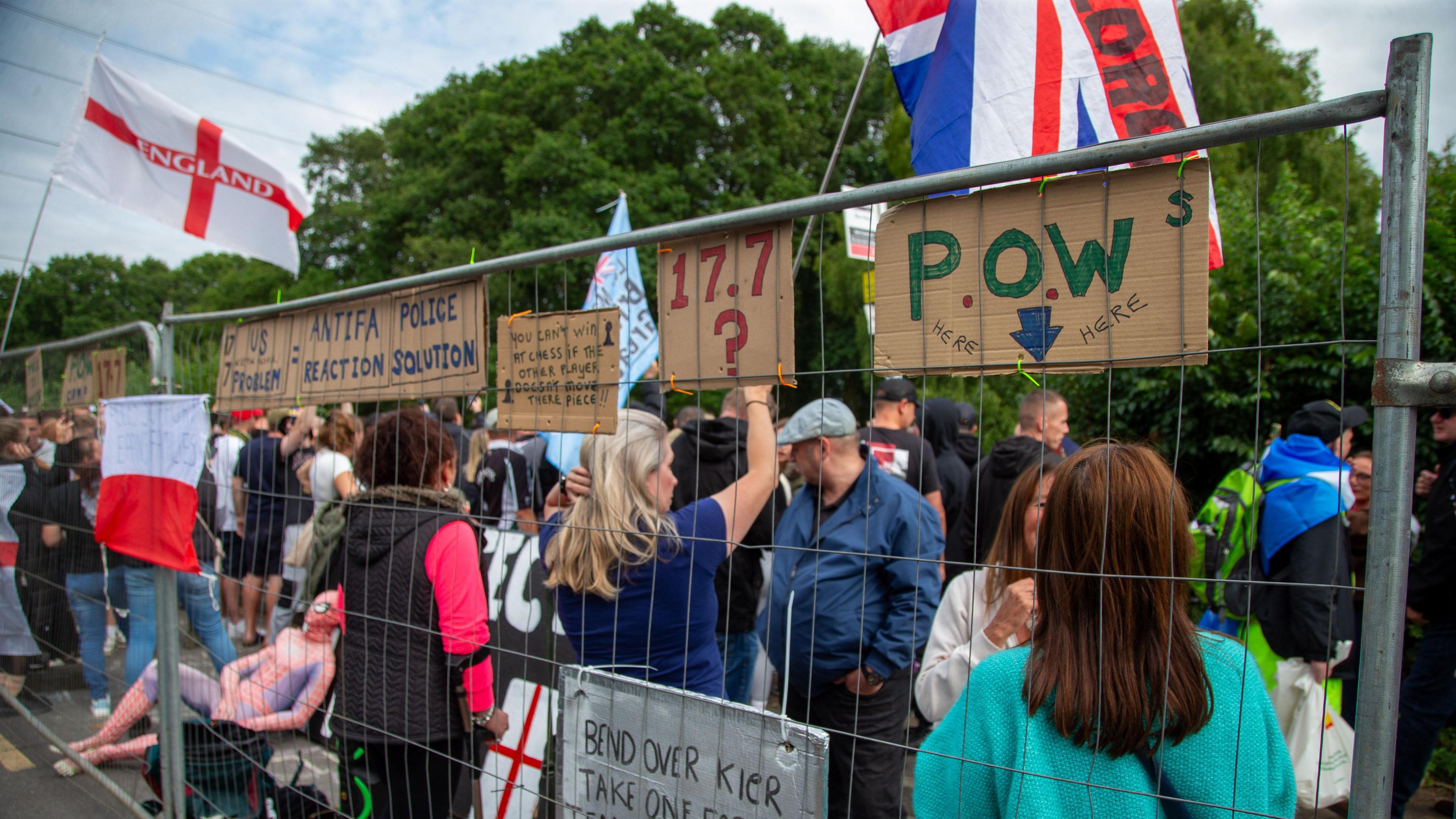 Anti-asylum seeker protesters some holding St George's cross and union flags standing behind a metal barrier.  