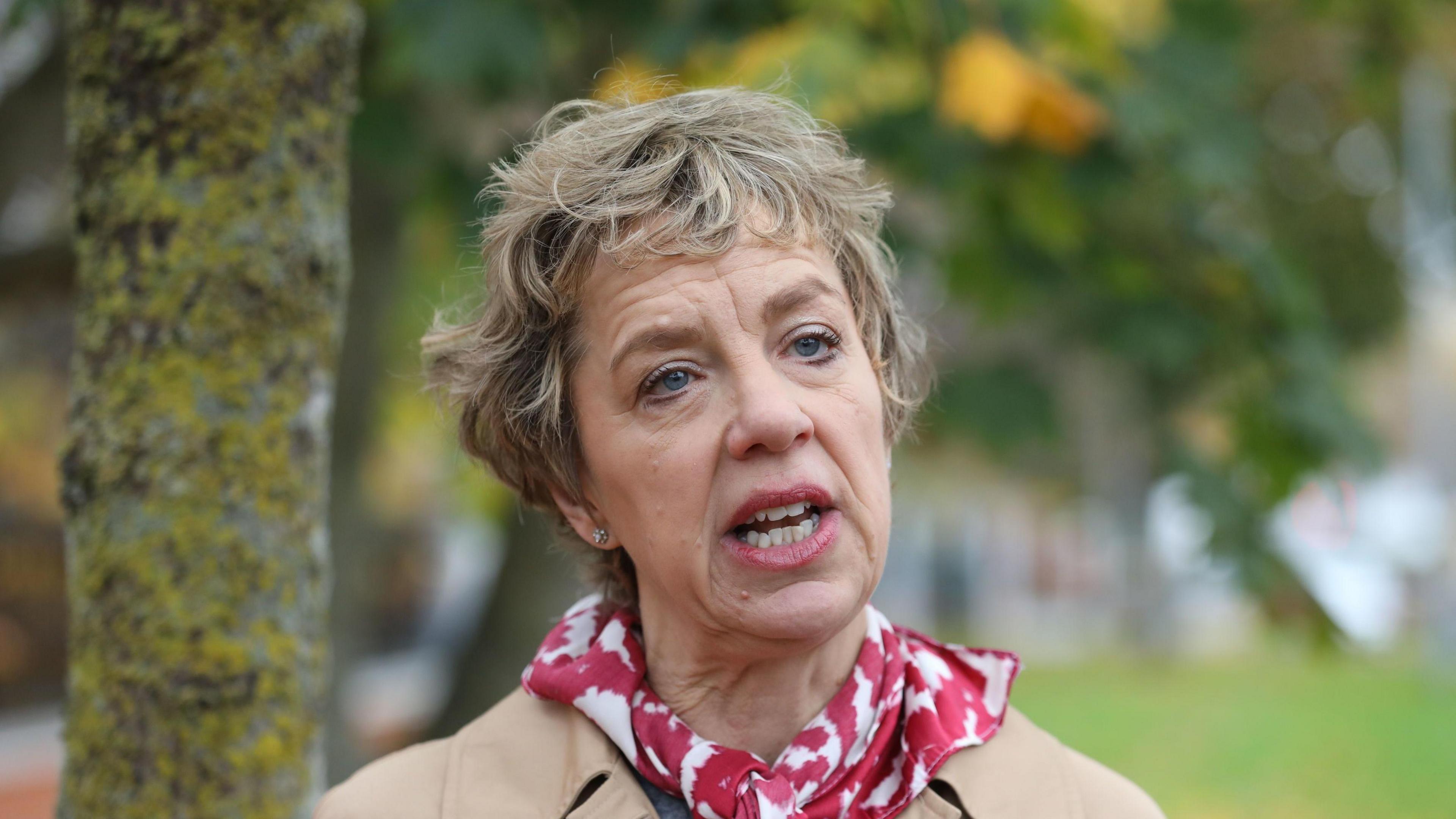 Ivana Bacik speaking to the media while canvassing in Kimmage, She has short light brown/grey hair. She is wearing a red and white scarf and beige coat. Trees can be seen in the background.