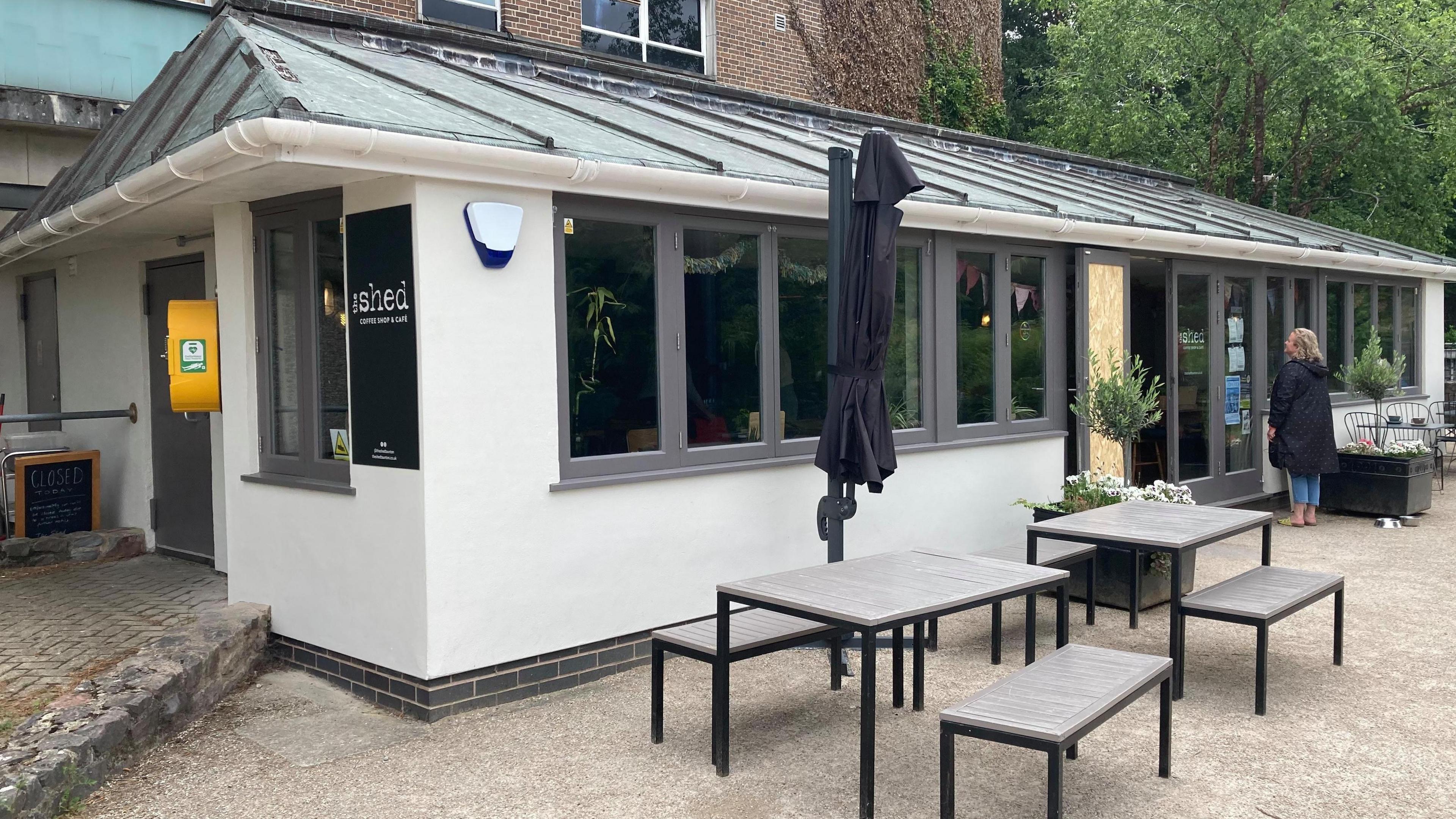 The picture shows the outdoor of The Shed. The walls are white and there are grey frames around the windows. There are benches and chairs outside.