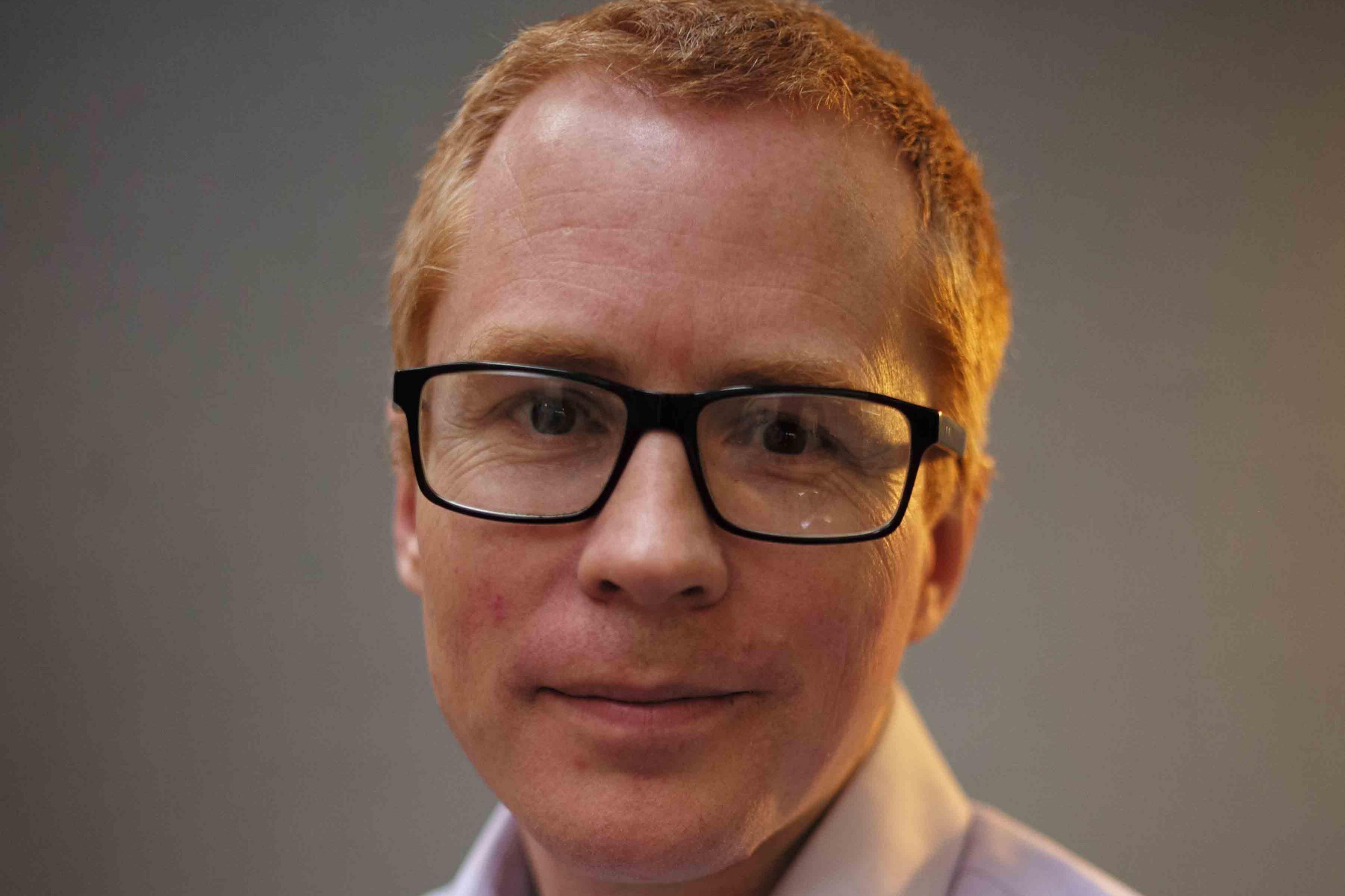 Jon Yates is smiling into the camera. He is wearing black-framed glasses and a purple shirt and has short ginger hair. He is in front of a grey background.