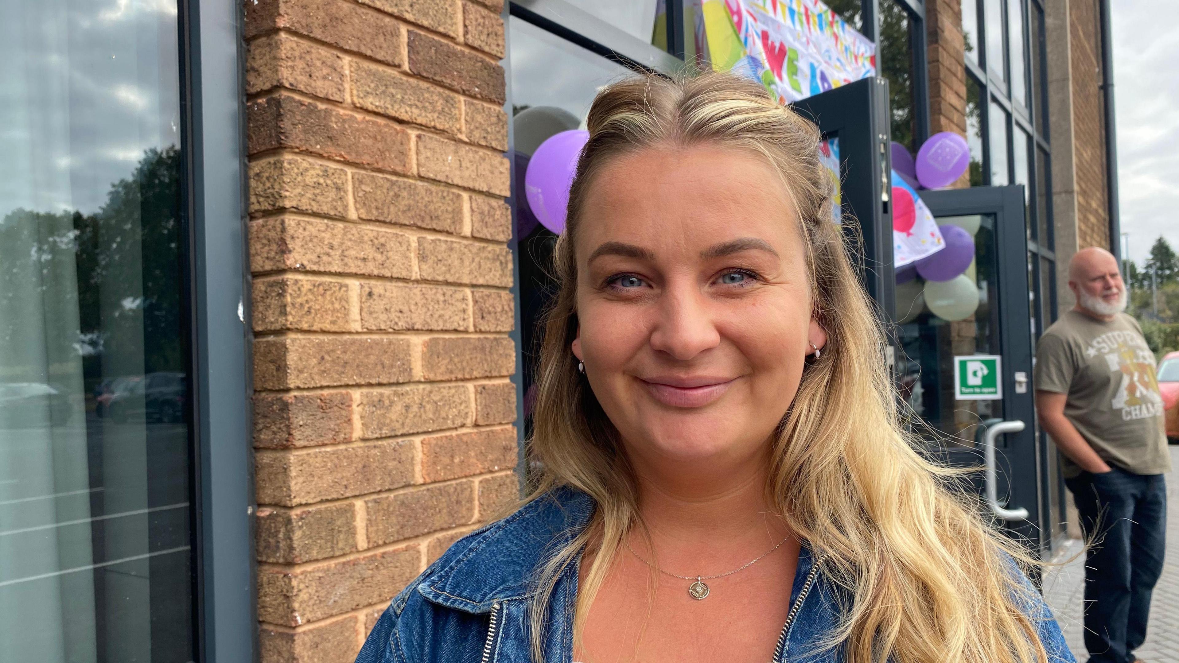 Ms Whitehouse looking into the camera and smiling. She has long blonde hair and is wearing a blue denim jacket. She's stood outside a school building, which is built of brown bricks and has large grey windows. Over the door, there's a rainbow banner reading 'Congratulations' and purple and white balloons are hung up. 