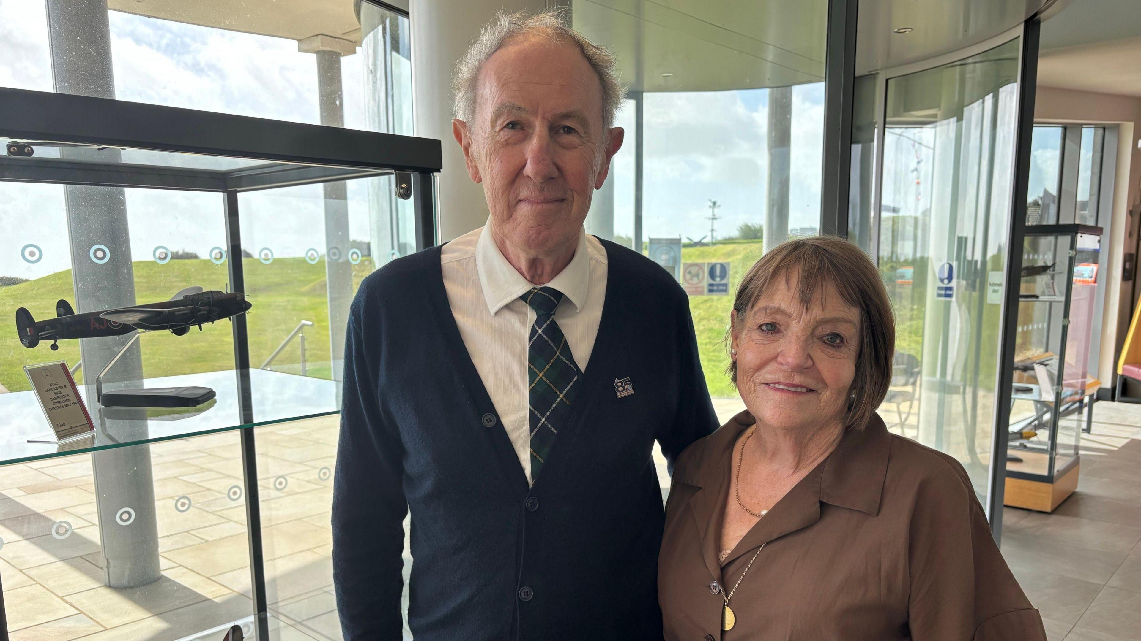 Alex Neill, the great grandson of Sir Keith Park, is standing next to Susie Hunt, who is the great granddaughter of Sir Keith. Mr Neill is wearing a black cardigan, a white shirt and a checked tie while Ms Hunt is wearing a dark brown shirt and two gold necklaces. They are inside a building featuring warplane models inside display cabinets. 
