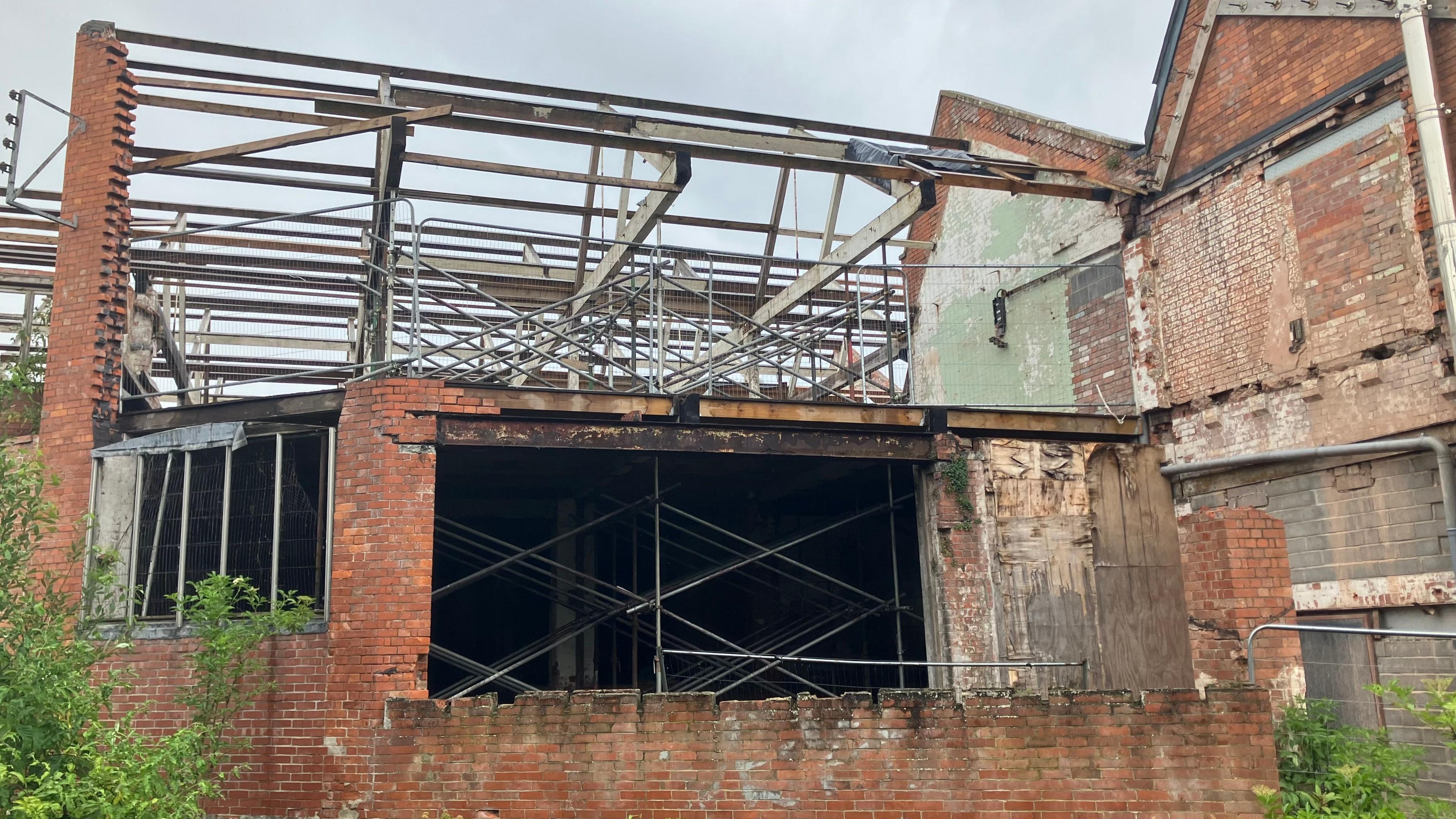 A derelict building with lots of exposed roofing beams and scaffolding that looks like it's supporting the inside of the building. Walls are completely missing in places.
