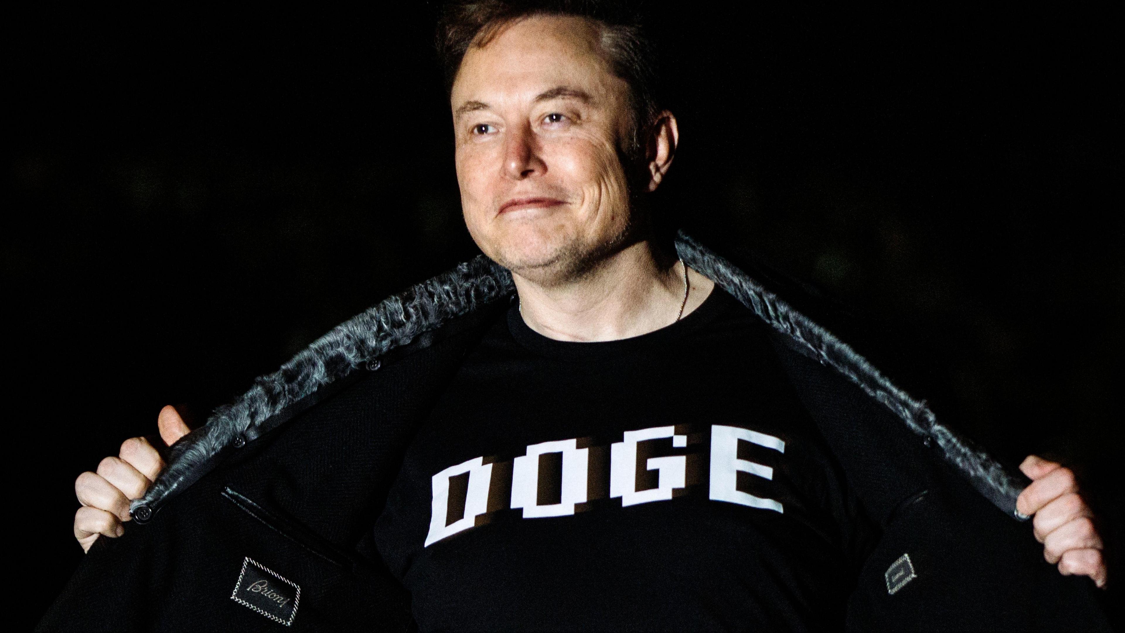 Elon Musk opens his jacket to reveal a t-shirt with the word DOGE in capitals written across his chest.