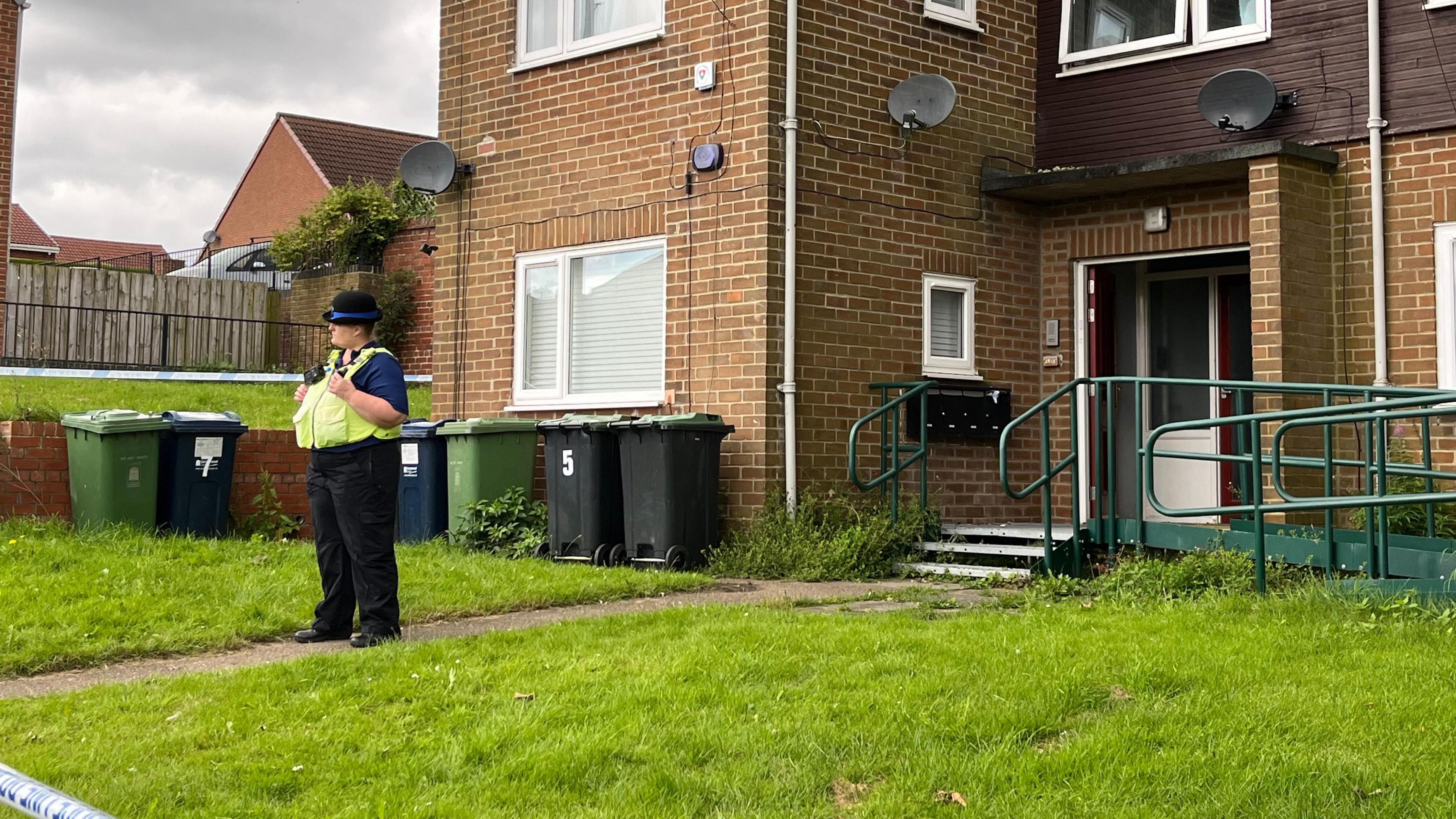 A female police officers standing in front of the property. Green steps and a metal ramp lead up to the front door and a number of wheelie bins are in the garden.