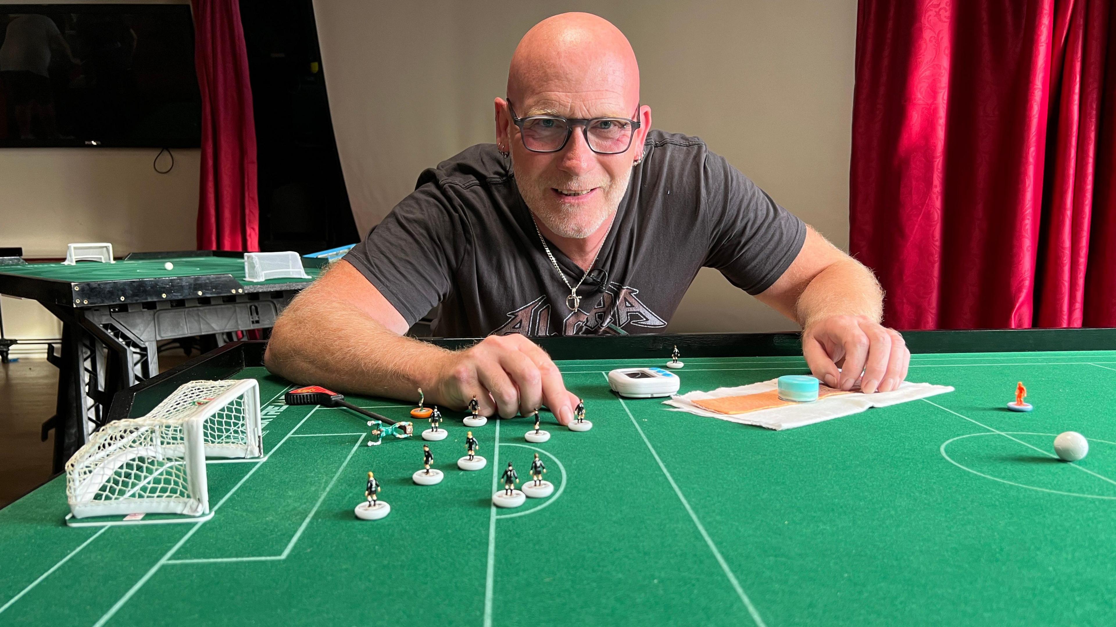 Mick Hammonds at table-top board with miniature players in front of him and red curtains in the background.