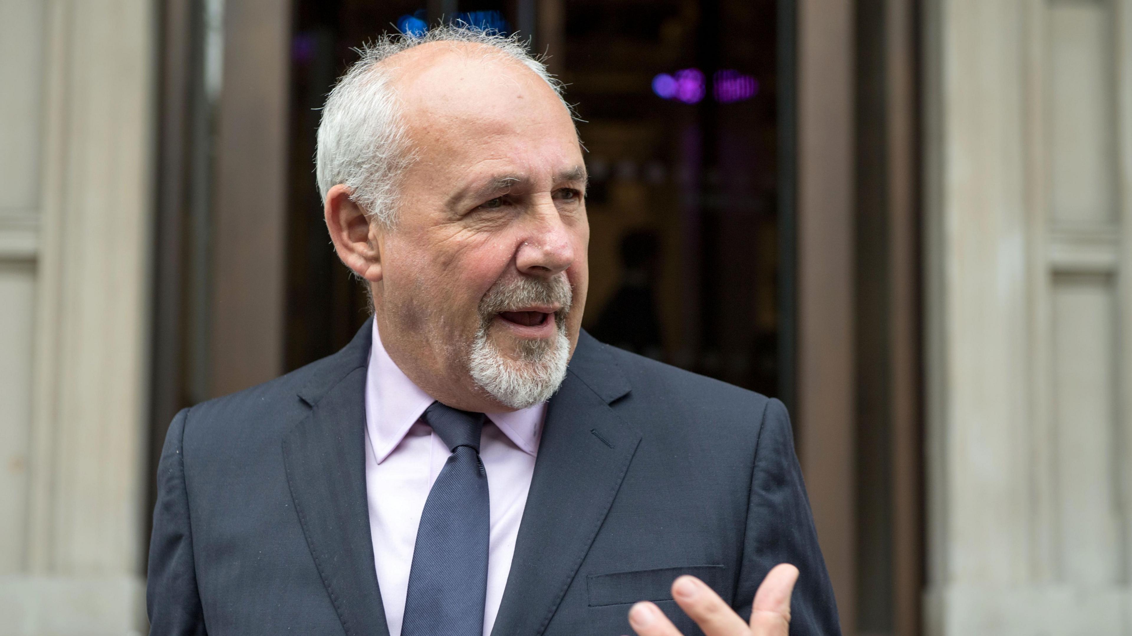 Labour MP Jon Trickett speaking outside a public building. He is a man with receding white hair, a goatee and he is wearing a dark suit with a lilac shirt