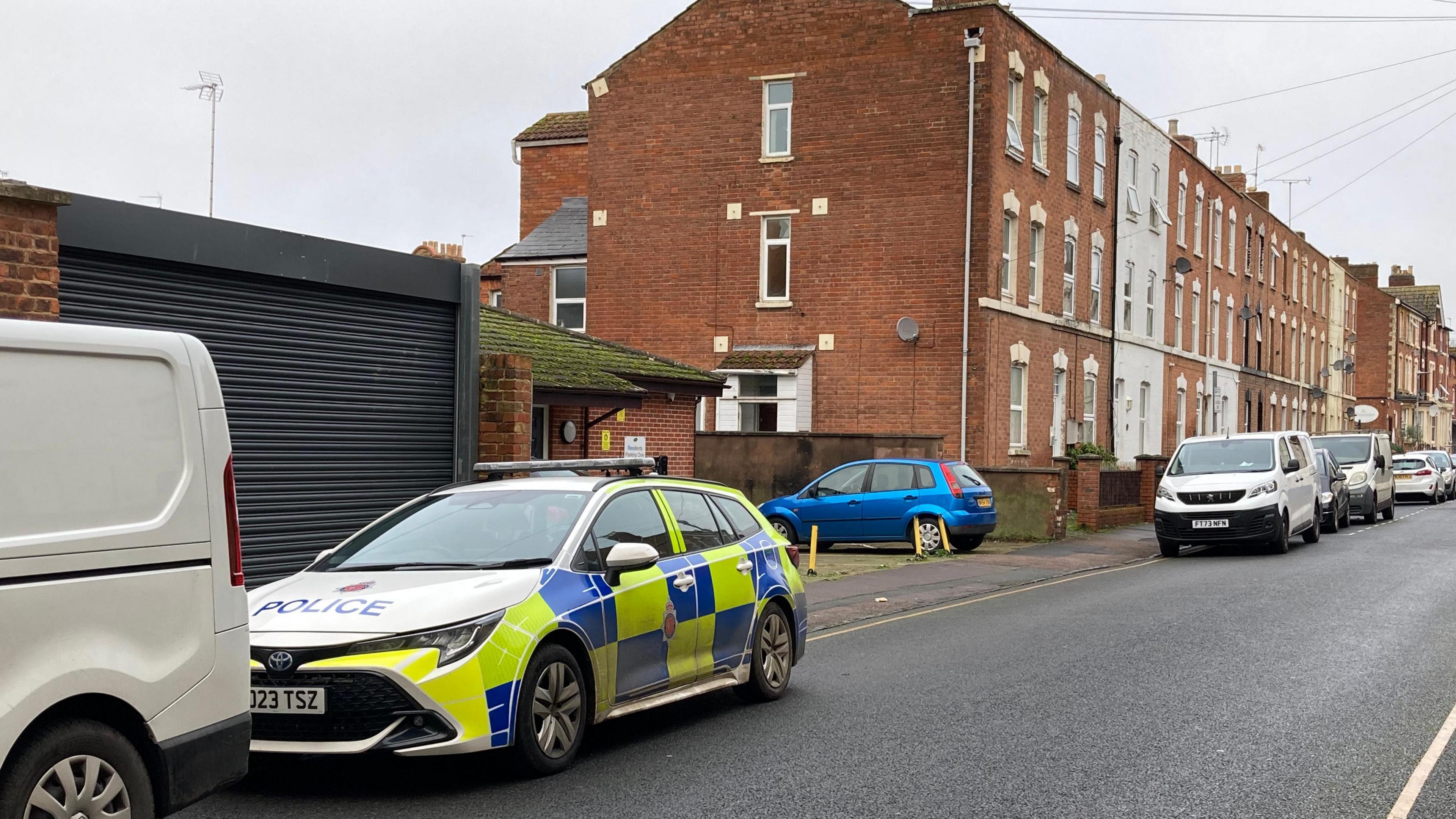 A police car in the foreground of a residential street with tall brick buildings lining the road