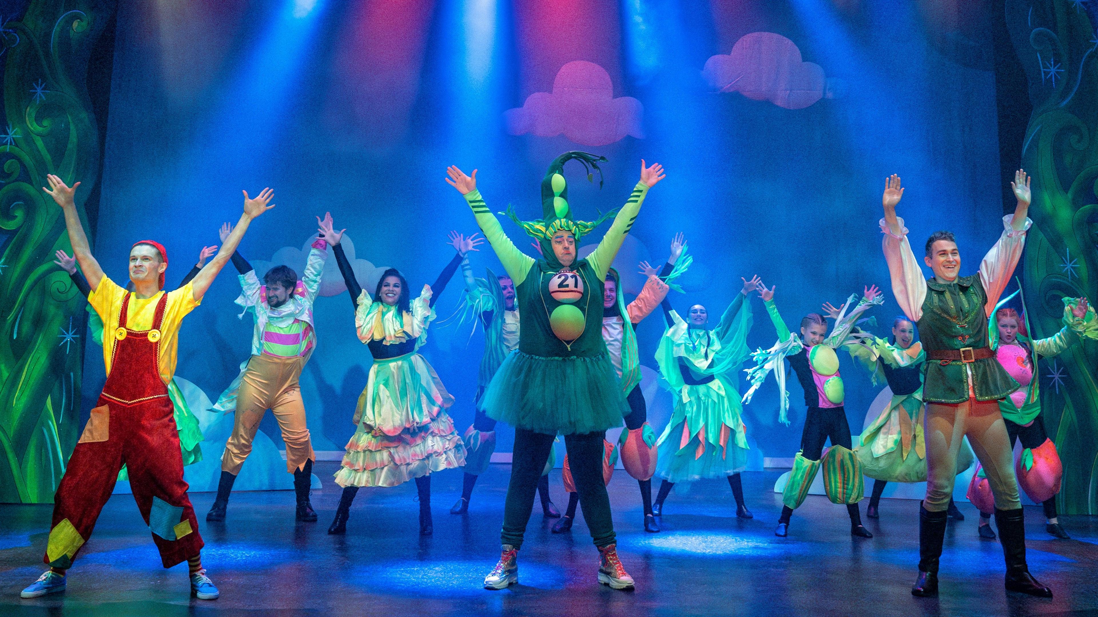 A stage with 11 people on. They all have their hands up and are stood as if they have just completed a dance number. At the front, a man is dressed as a pea pod, with large sponge peas stuck in a row up his chest up to the top of his head. Around him are people wearing bright colours and smiling widely. To his right is a man who appears to be playing jack, wearing black boots and green trousers and a top with large white sleeves. The stage is lit in blue light.