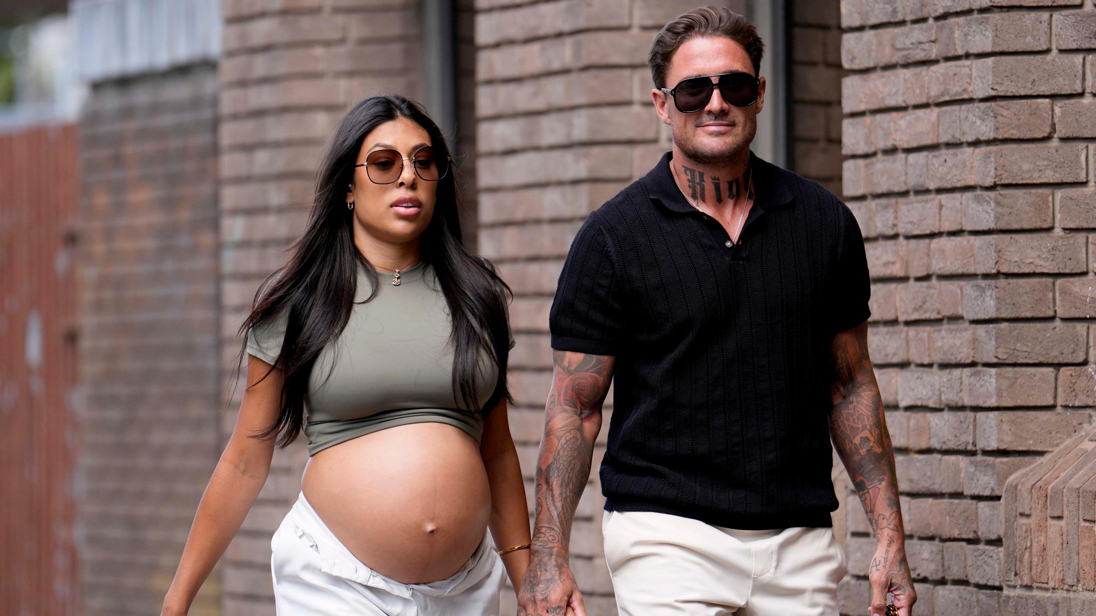 Stephen Bear (right) holding hands with Miami (left) as they walk into court, with a brick wall behind them. Miami is heavily pregnant with her bump showing and she has long black hair and big sunglasses on, and white trousers.