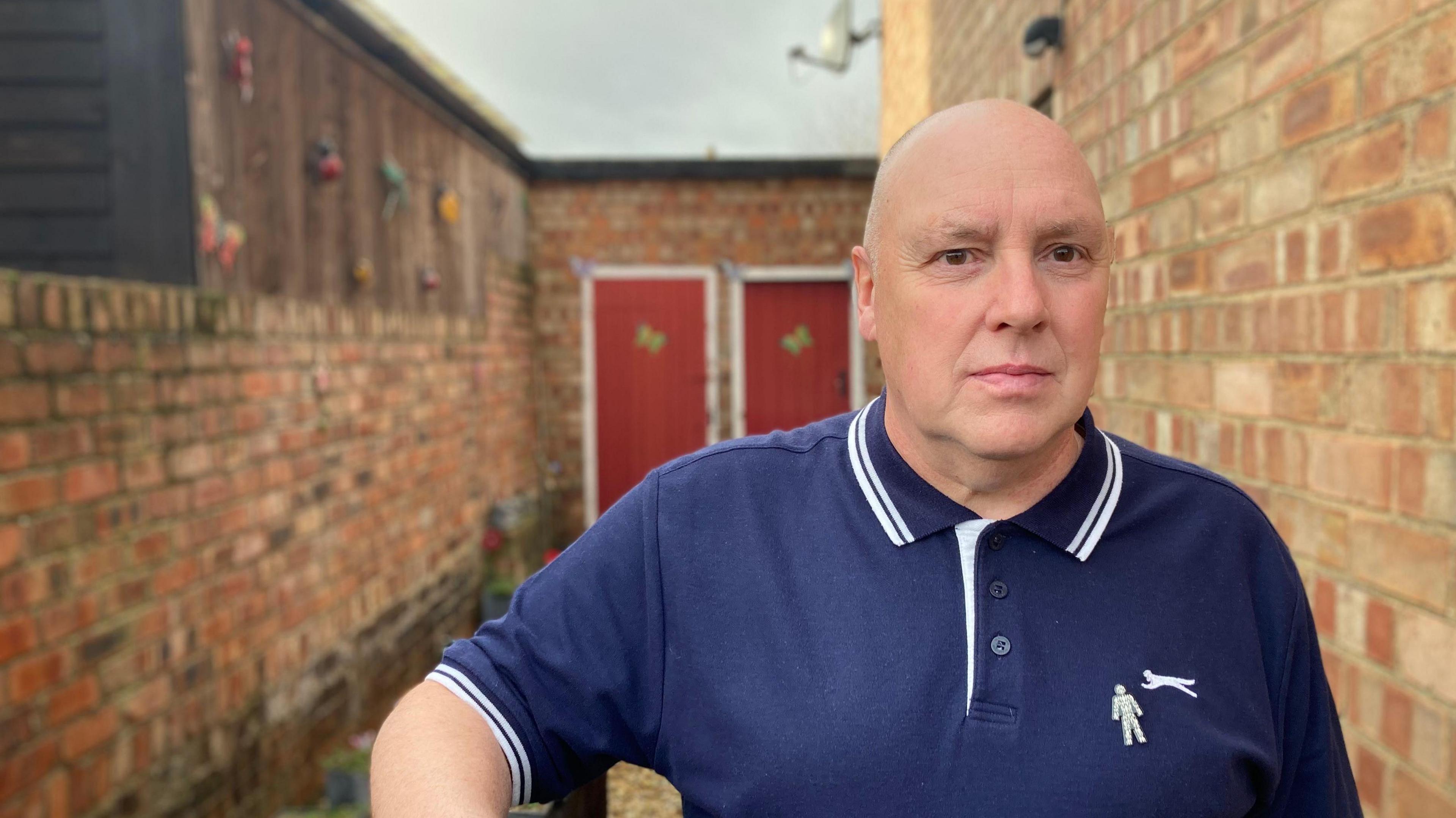 A man with a shaved head, dressed in a blue polo shirt, stands in a narrow yard between two red-brick walls. Two red wooden doors with white frames can be seen in the background.