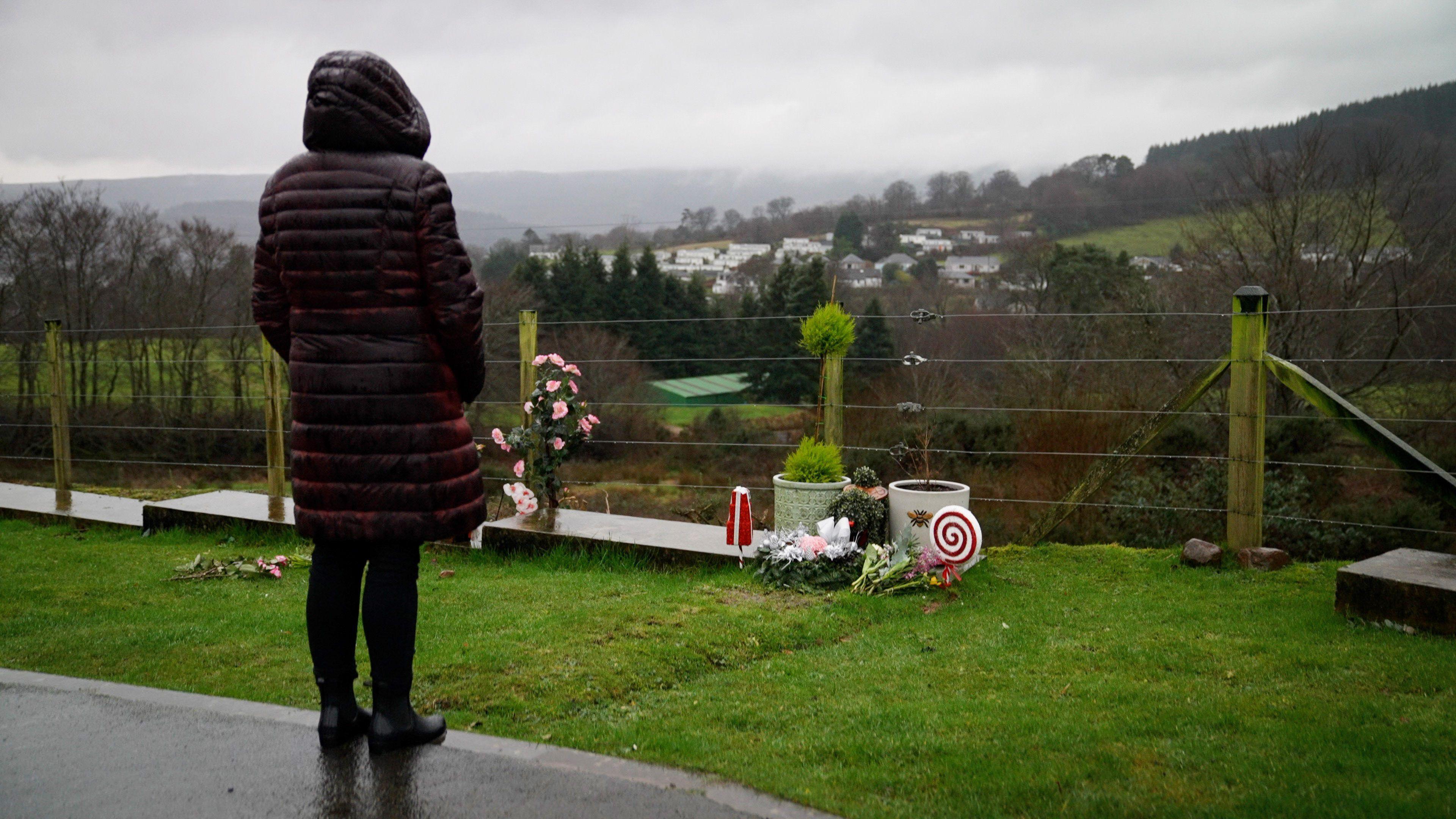 A woman in a burgundy jacket, with her hood up, looking at a grave on the edge of a cemetery. In the background is a view of trees, fields and houses.