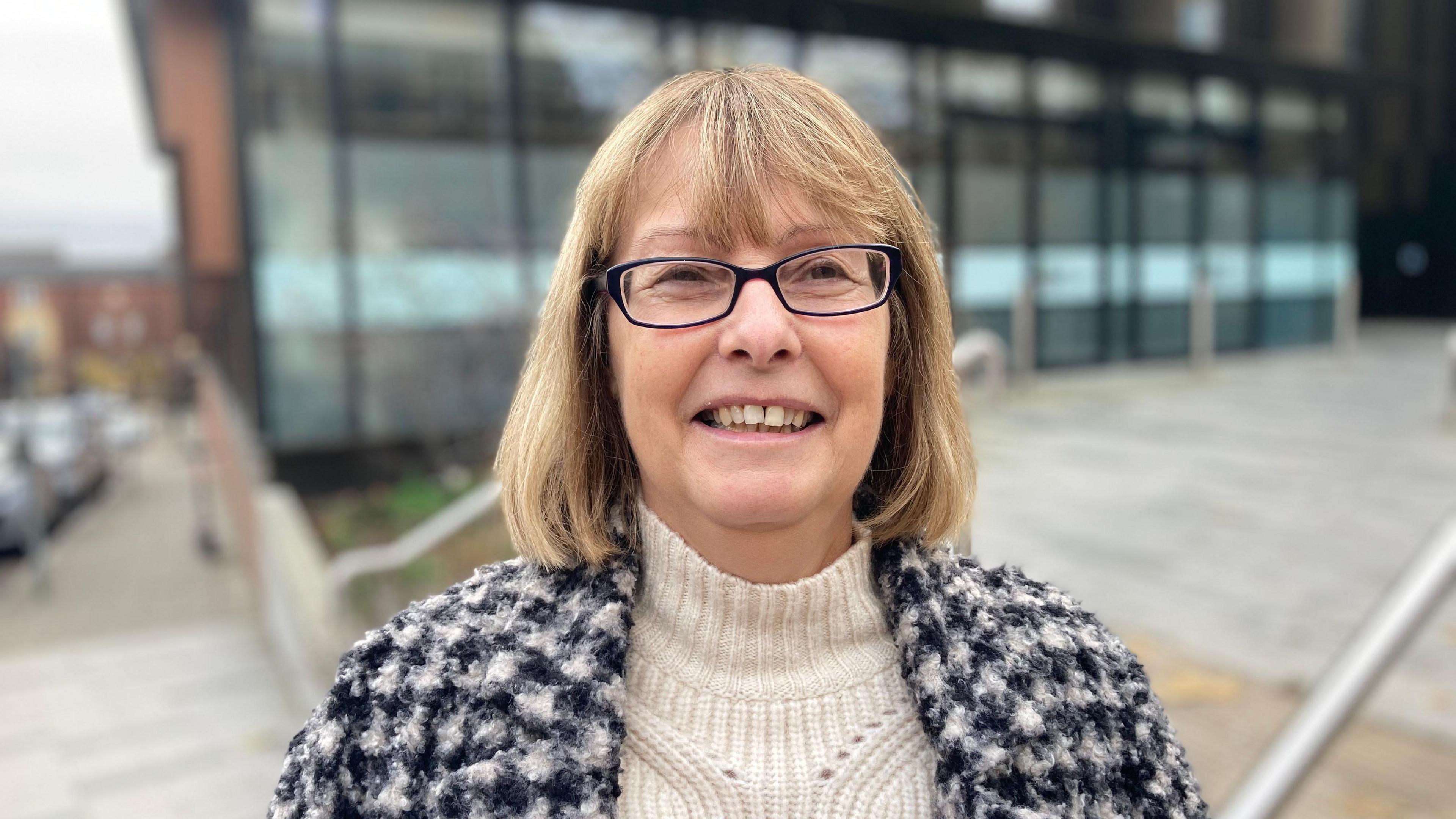 A woman with glasses in a black and white knitted top over a cream funnel neck jumper stands on some stairs looking at the camera. 